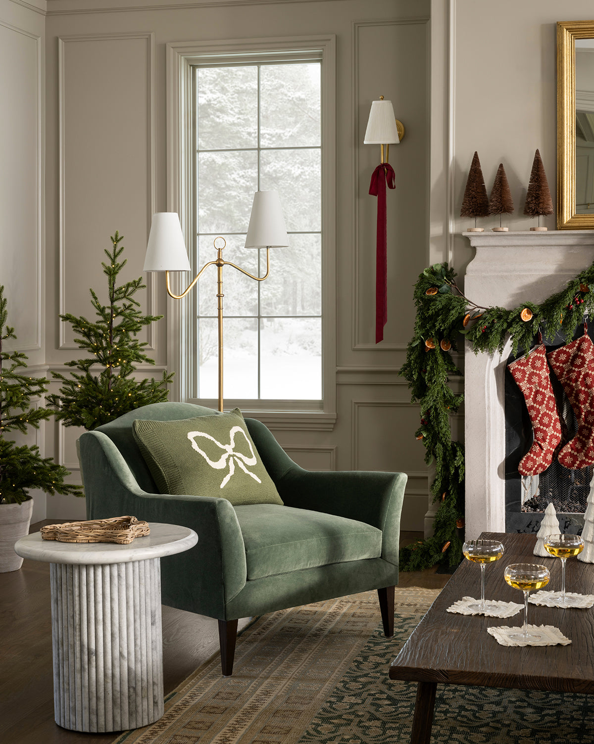 A cozy living room with a green armchair, the Bella Pillow Cover featuring a white bow, knit accents, a side table, decorated Christmas trees, garland on the fireplace, stockings hanging, and snowy scenery outside the window, by Vendixs