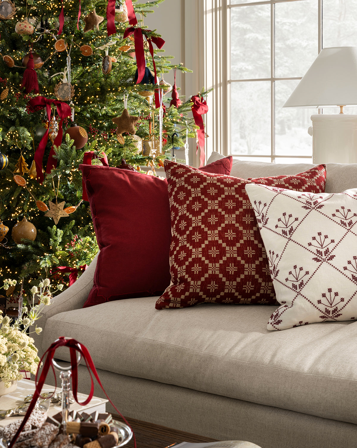 A beige sofa with McCallister Pillow Covers and red-and-white patterned pillows sits by a festive Christmas tree. Sunlight fills the room, highlighting holiday decor and treats arranged on the table in front, by Vendixs
