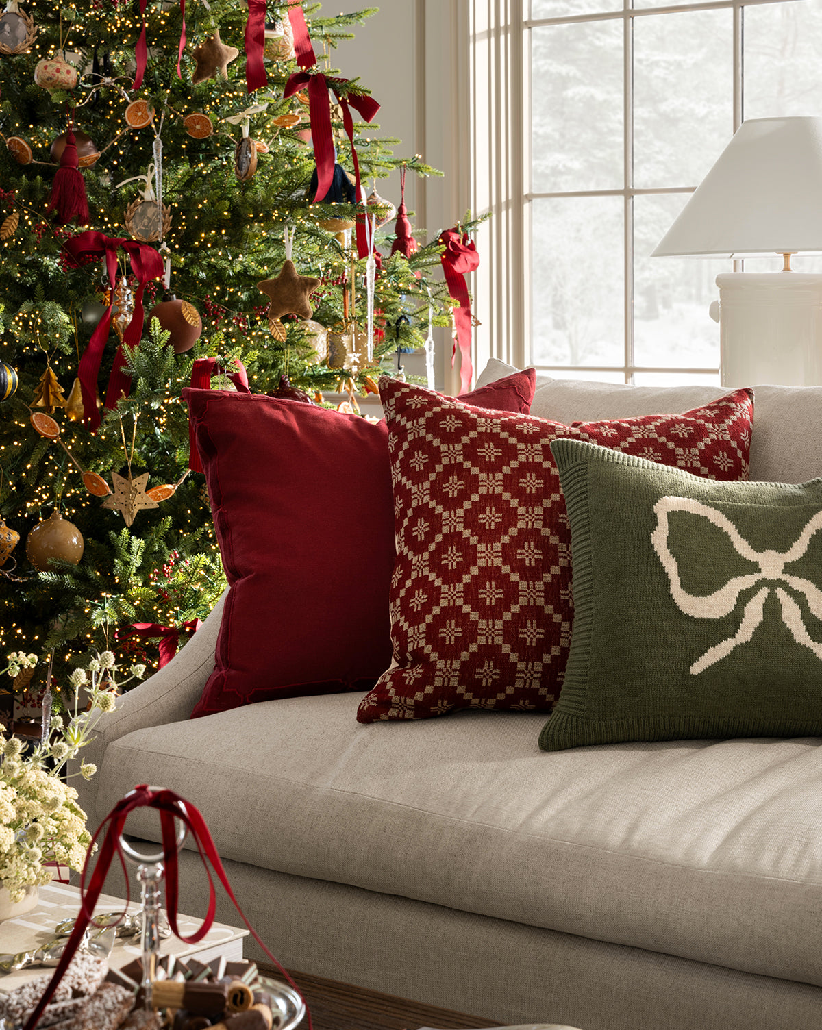 A cozy living room with seasonal styling features a decorated Christmas tree, Jude Pillow Covers in red and green on a light-colored sofa, and a window framing a snowy landscape outside, by Vendixs