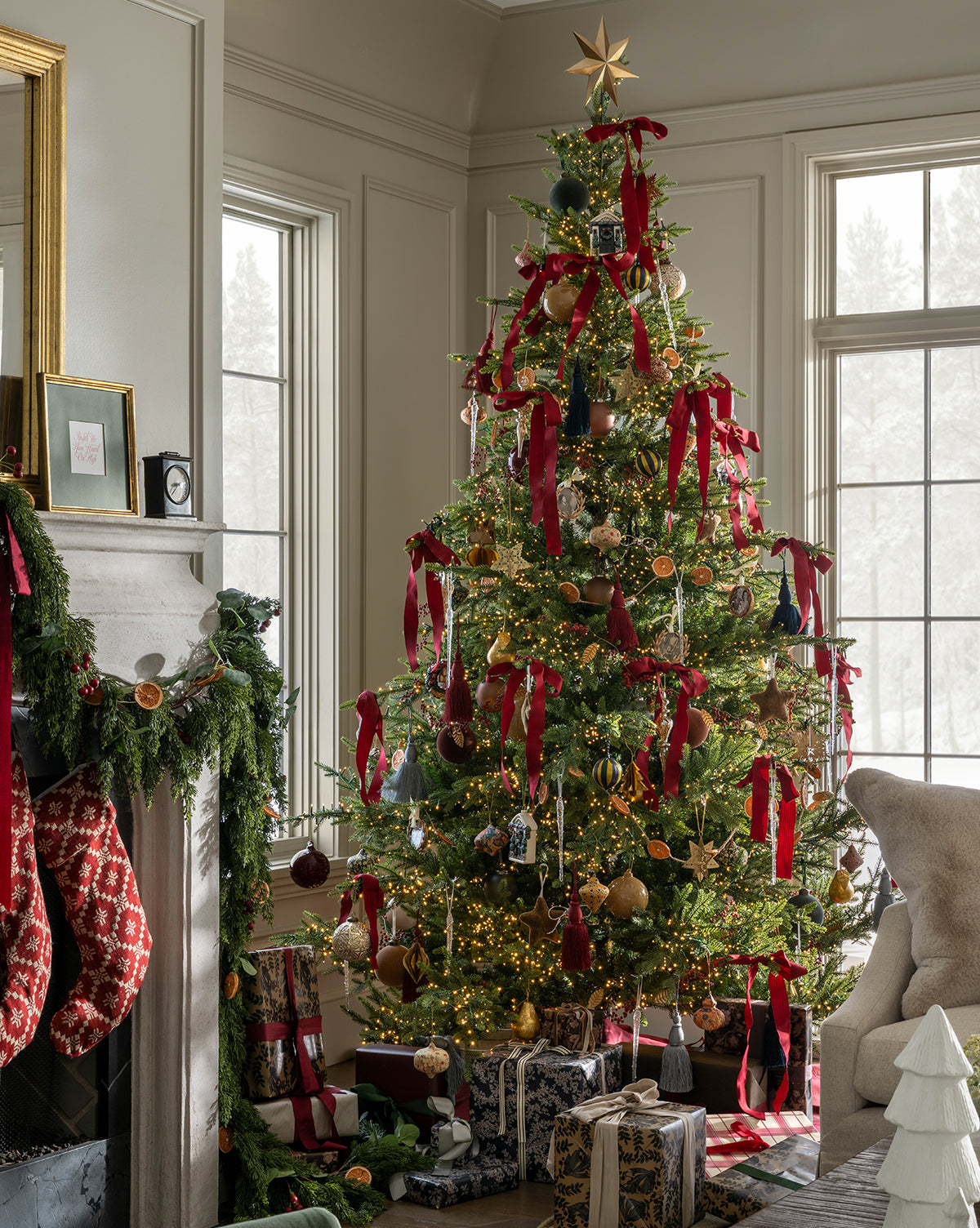 The Classic Christmas Tree, inspired by the Grandis Fir, stands by the window decorated with red ribbons, ornaments, and a gold star. Below rest presents; nearby, stockings and garland adorn the mantel in a sunlit living room, by Vendixs