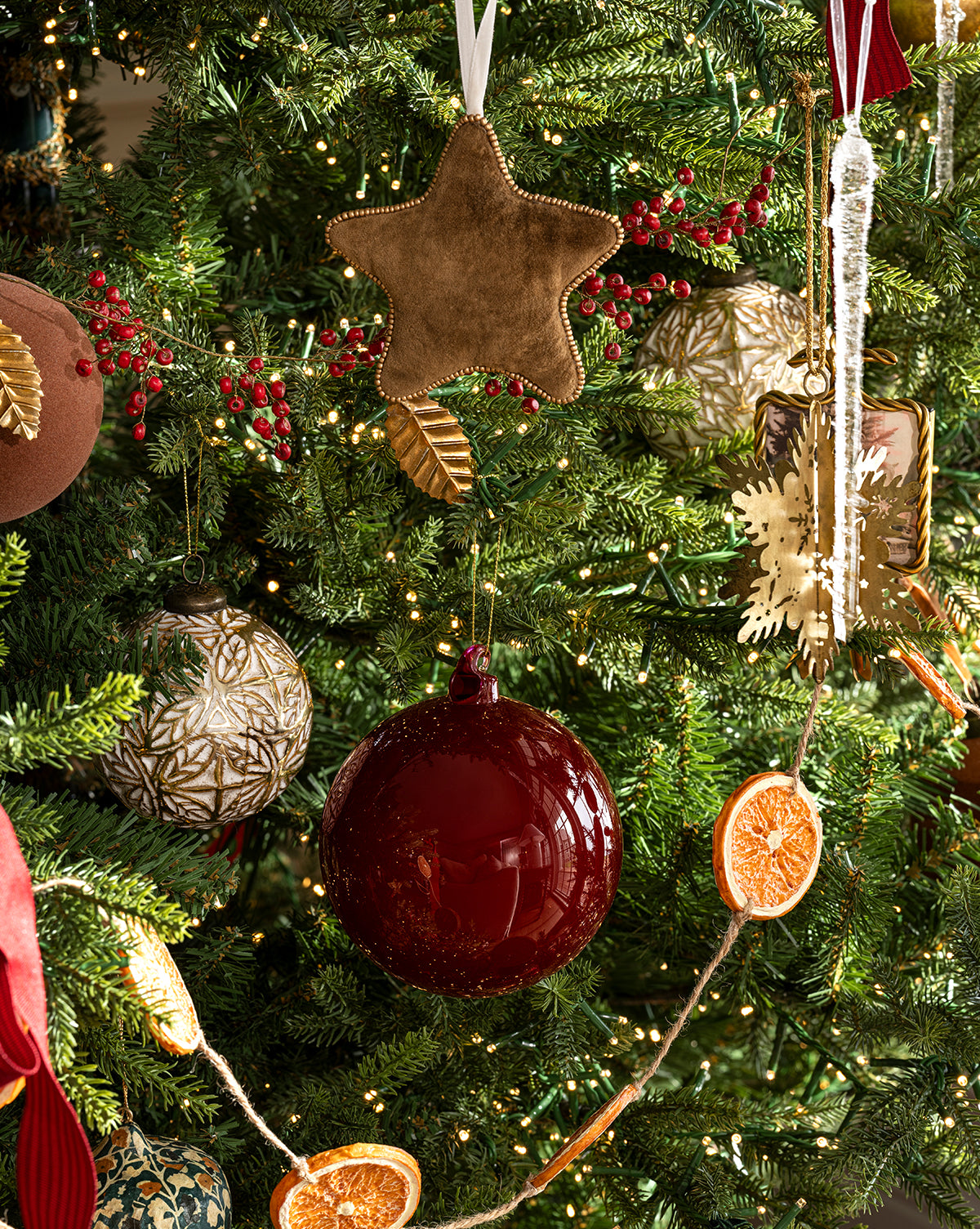 This holiday tree glows with gold and red ornaments, a Plush Beaded Star Ornament, dried orange slices, a gold snowflake, twinkling white lights, green branches, and red berries, by Vendixs