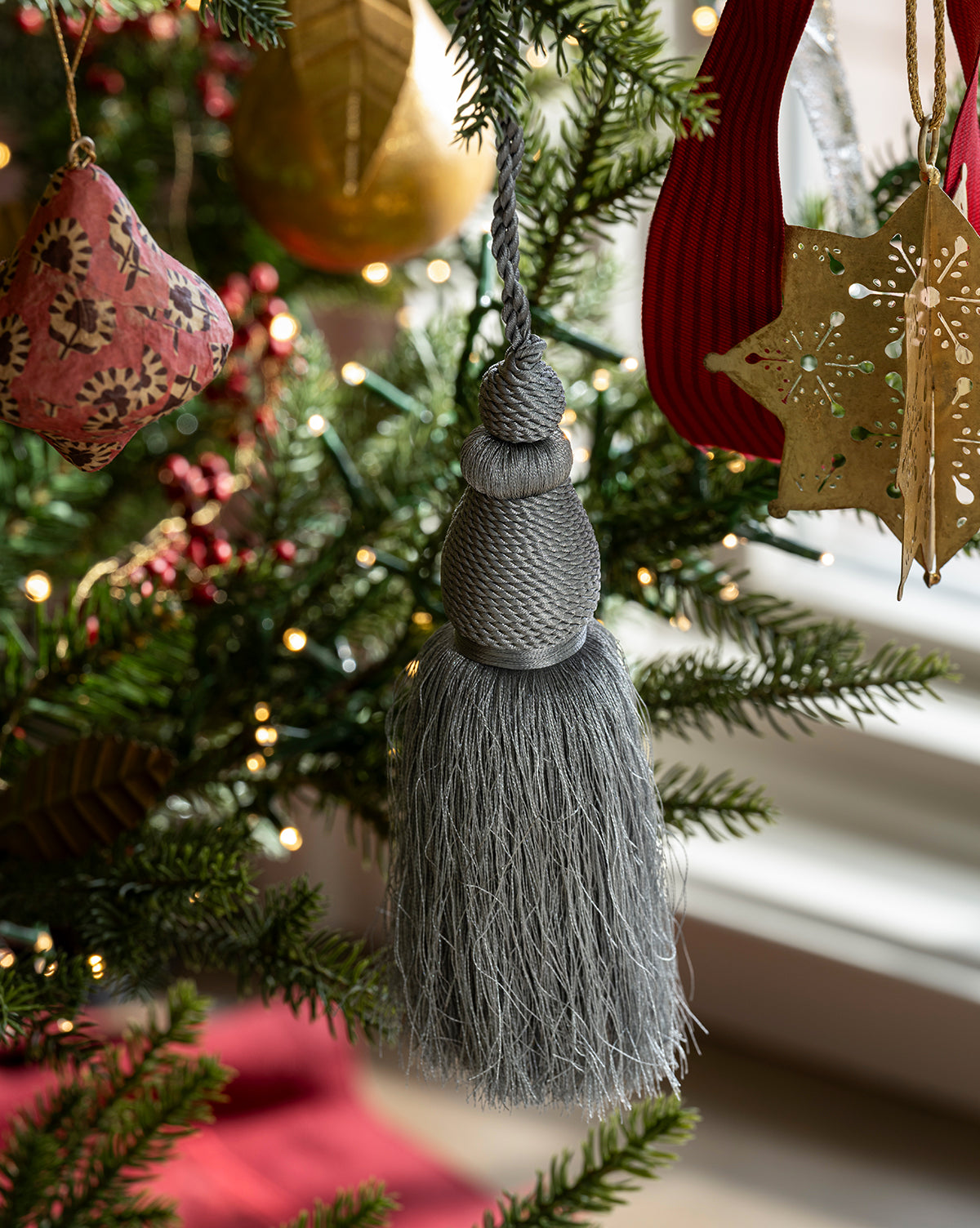 The Lucia Bullion Tassel hangs on a festive Christmas tree with green branches, twinkling lights, and ornaments like a gold star and red fabric shape, bringing vintage-inspired charm to your holiday décor, by Vendixs