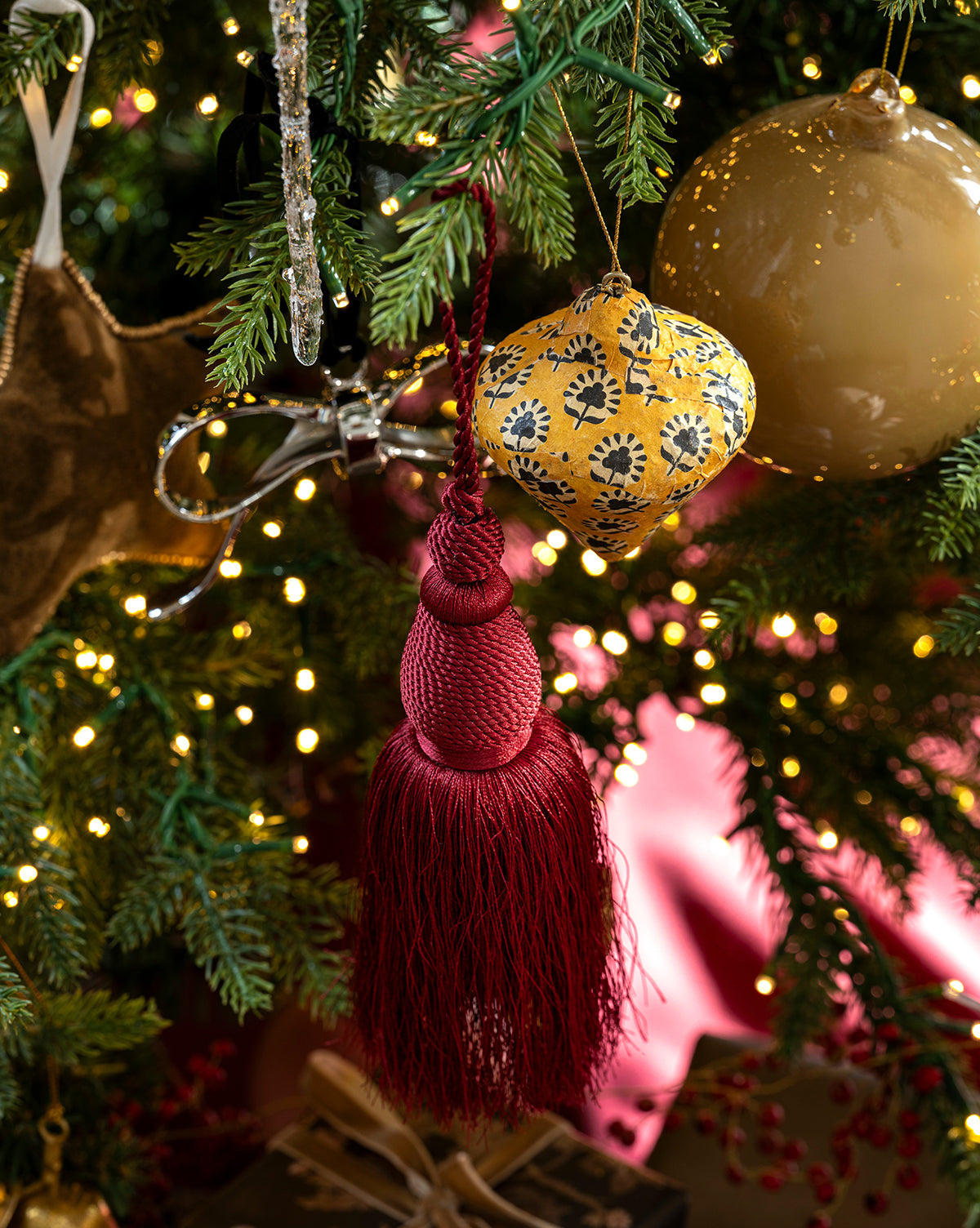 A close-up of a decorated Christmas tree featuring the maroon Lucia Bullion Tassel ornament, a yellow patterned bauble, gold ornaments, twinkling fairy lights, green pine branches, and a wrapped gift at the base, by Vendixs