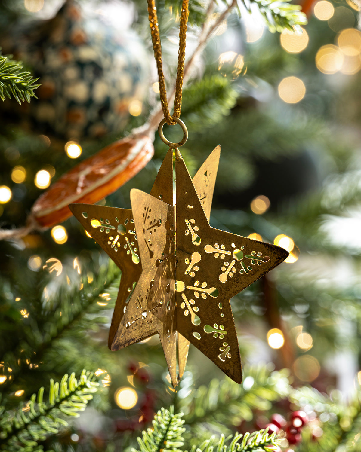 A gold, star-shaped metal ornament with cut-out designs hangs from a Classic Christmas Tree branch, surrounded by lush green needles and softly blurred twinkling lights in the background, by Vendixs