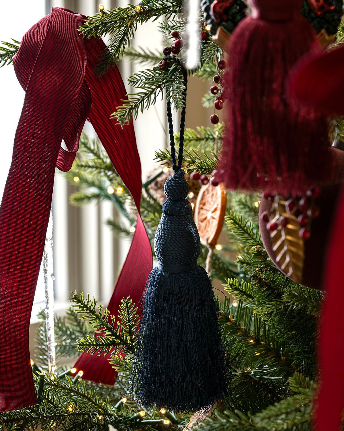 A close-up of a Christmas tree decorated with a dark blue tassel ornament, dried orange slices, small lights, and a 1.5" Woven Grosgrain Stripe Ribbon with Woven Edge among lush green pine branches, by Vendixs