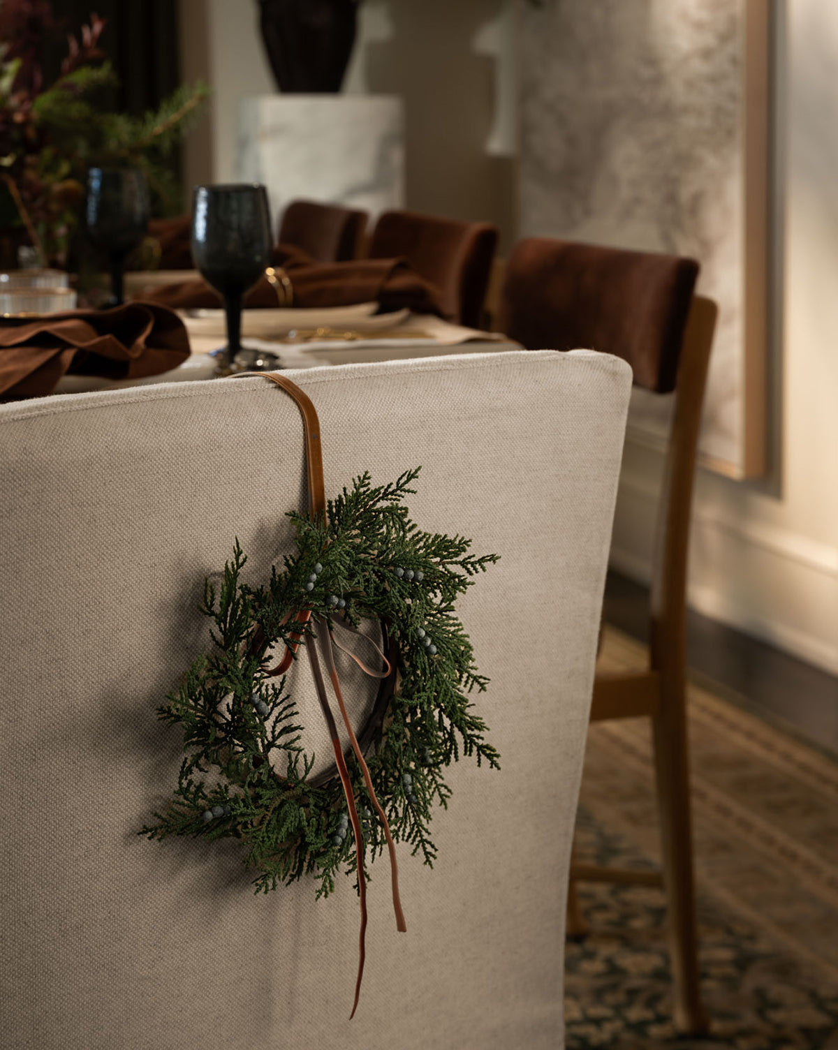 An 8" Blueberry & Juniper Faux Wreath with ribbons hangs on the back of a beige dining chair at a table set with dark glassware and brown napkins, creating an elegant, festive holiday atmosphere, by Vendixs