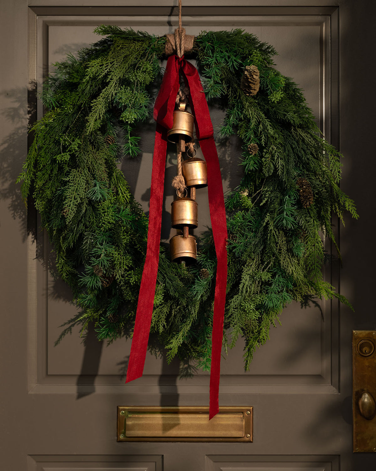A green holiday wreath with pinecones and a red ribbon hangs on a gray door. Hanging Iron Bells with a gold finish add to the festive décor, while a brass mail slot is visible below, by Vendixs