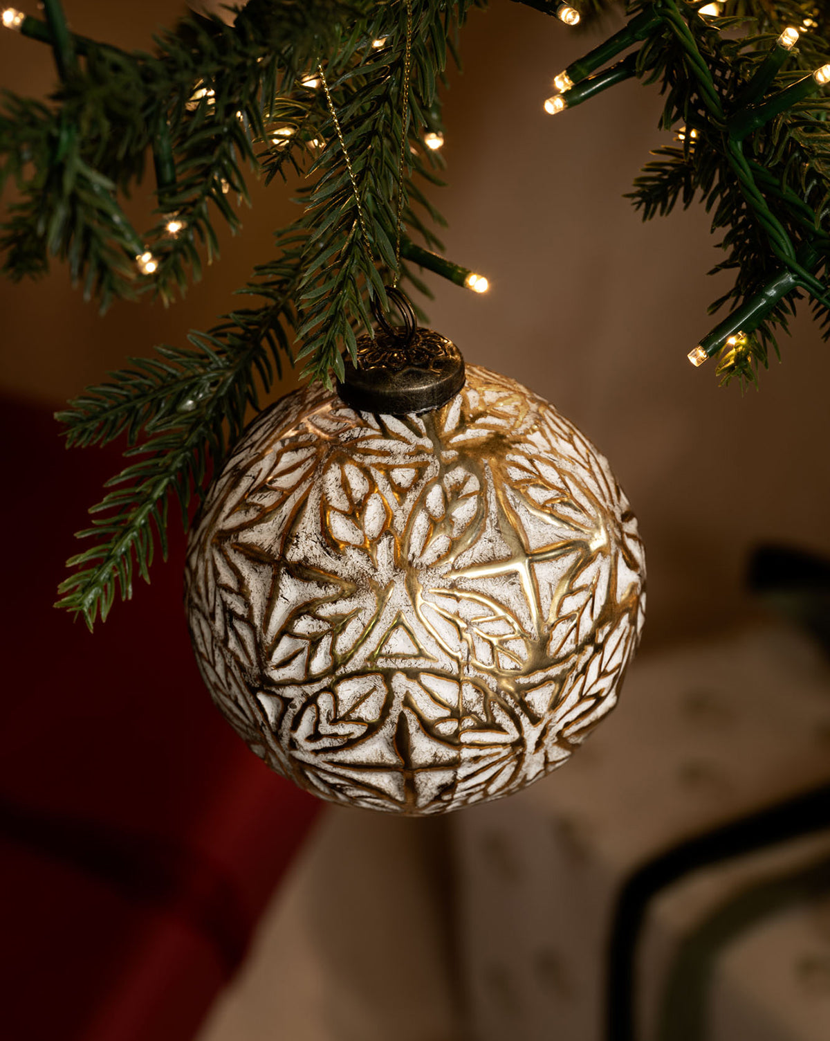 The Alessandro Glass Ornament, featuring a white and gold motif, hangs from a lit evergreen branch with warm string lights and blurred holiday decor in the background, by Vendixs