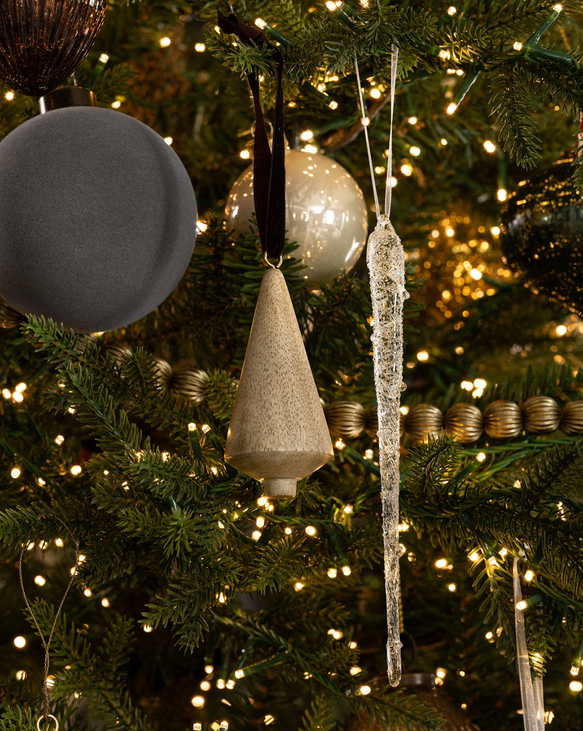 A Christmas tree adorned with elegant decorations, including a matte gray ball, a wooden cone ornament, a set of 3 Frosted Icicle Ornaments, gold bead garland, and twinkling string lights, by Vendixs