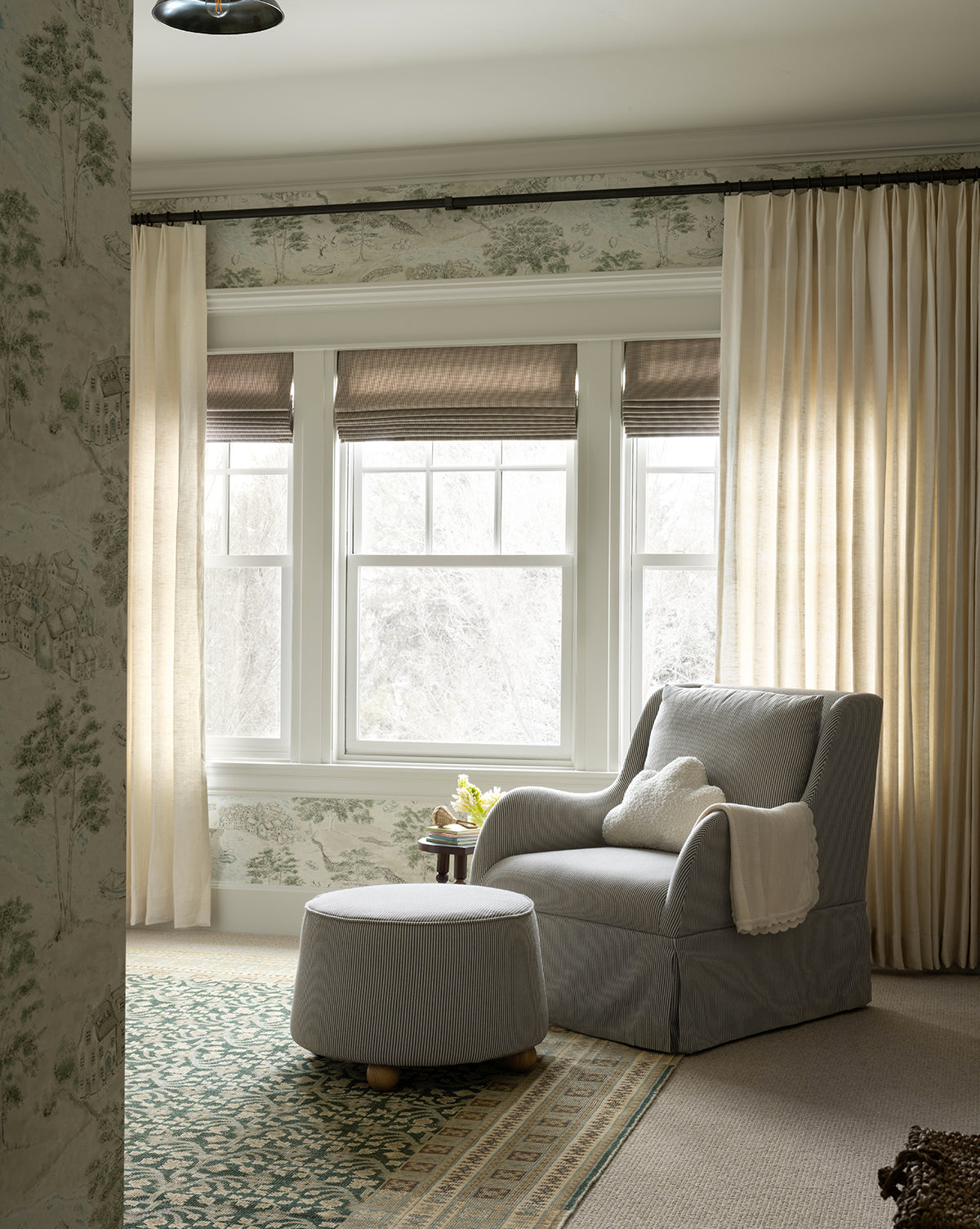 A cozy reading nook with a plush seat, Barnaby Ottoman in gray, a cream throw blanket, green patterned rug, ash wood accents, sheer curtains, and large windows that fill the space with soft natural light, by McGee & Co.