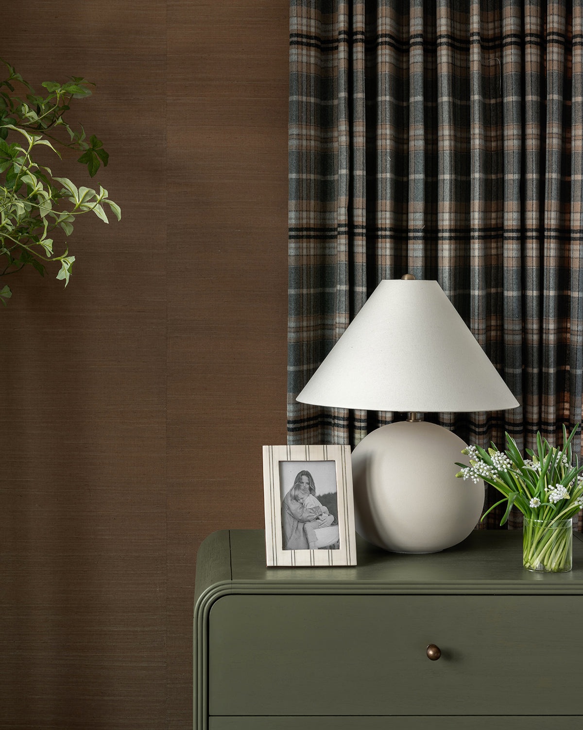 A green dresser topped with the Tess Ceramic Table Lamp, a framed black-and-white photo, and a small vase of flowers stands before plaid curtains and a brown wall. Green plant leaves hang in the upper left corner, by Vendixs