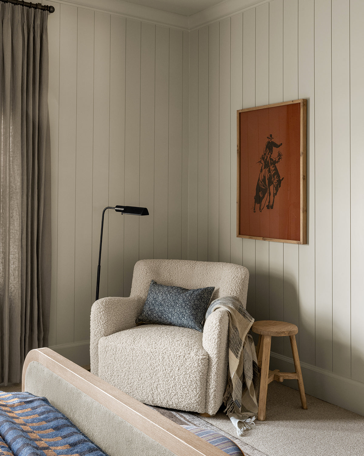 A cozy reading nook features the Magda Lounge Chair (Ready to Ship) with a blue-gray pillow and plaid throw blanket. A wooden stool and black floor lamp are nearby; Western art on white shiplap walls, with gray curtains framing the window, by Vendixs