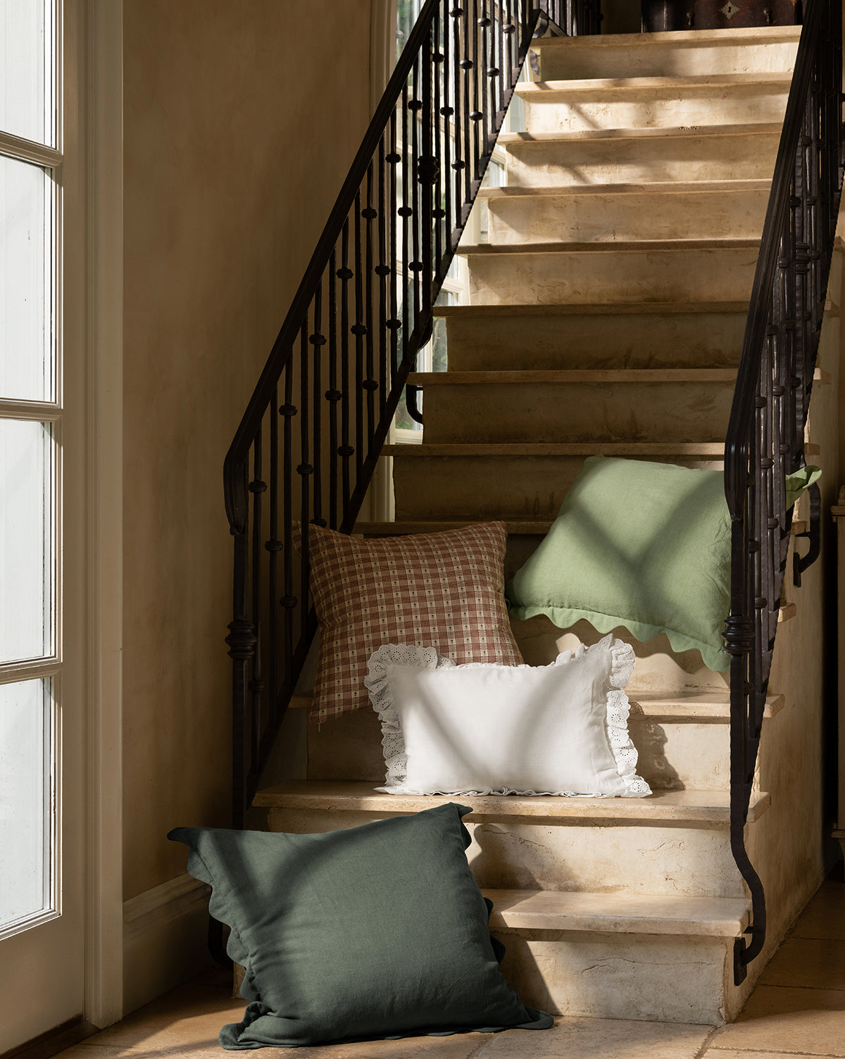 A sunlit stone staircase with a black iron railing is decorated with Vendixs x Loeffler Randall Colette Scallop Border Pillow Cover, plus green, ruffled white, and pink plaid pillows. Light from the left window highlights their cozy mix of textures, by Vendixs