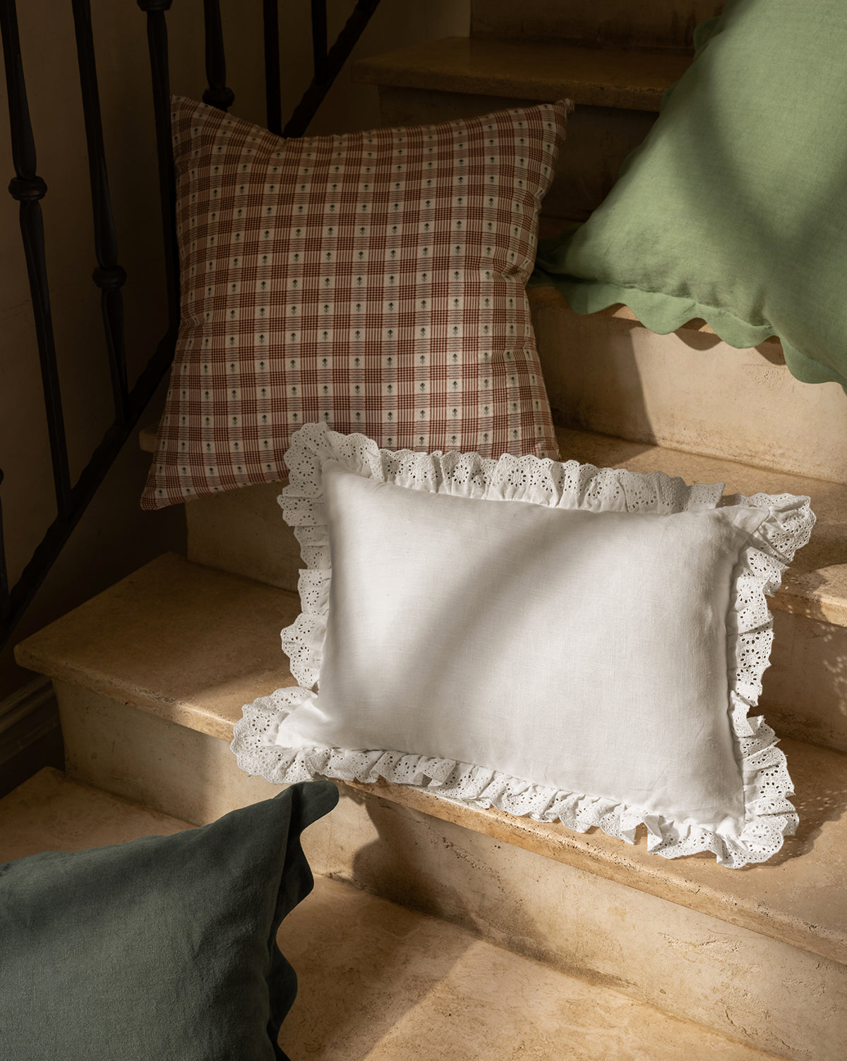 Three decorative pillows rest on stone steps: a green pillow with a wavy edge, a brown checkered cushion, and the Vendixs x Loeffler Randall Delilah Broderie Trim Pillow Cover in white linen. Sunlight casts soft shadows across the scene by Vendixs