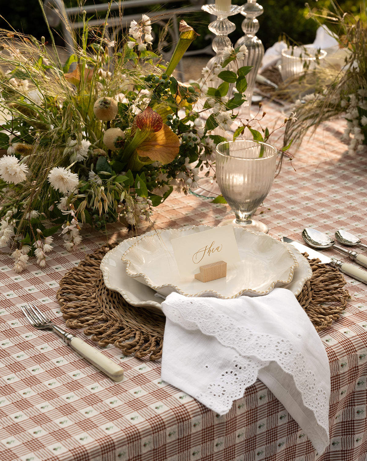 A rustic outdoor table set features a checked tablecloth, woven placemat, scalloped plate, Vendixs x Loeffler Randall Delilah Broderie Napkins (Set of 4), glassware, silver cutlery, a name card, and wildflowers in natural light by Vendixs
