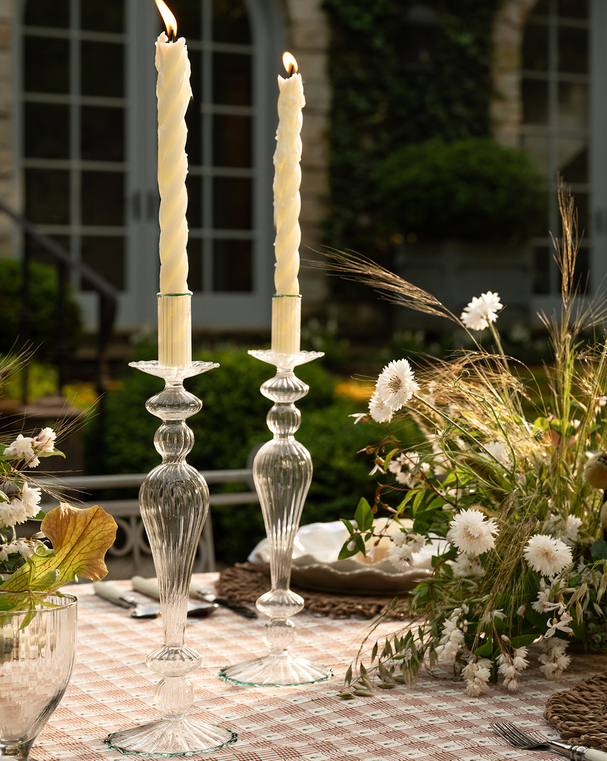 Two tall ivory candles in Vendixs x Loeffler Randall Effie Taper Holders sit on a checkered tablecloth, surrounded by white and beige dried flowers and greenery, with a blurred garden and building in the background, by Vendixs