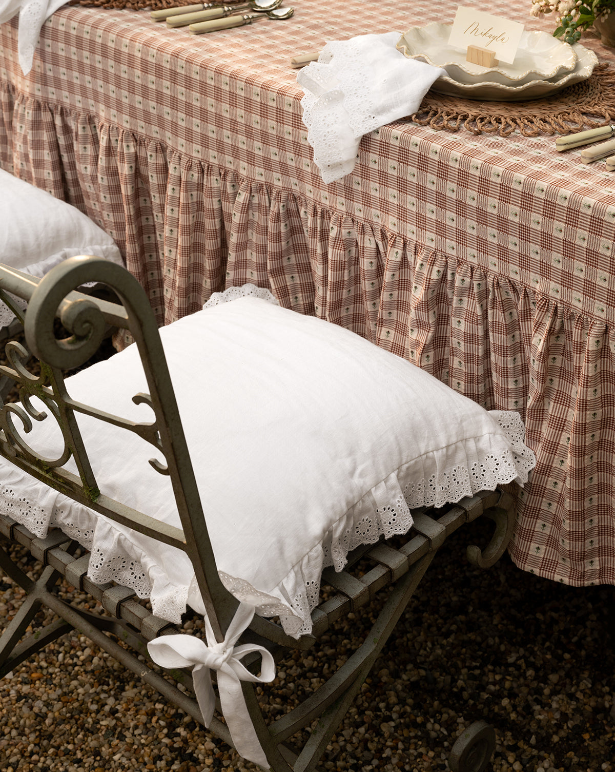 A vintage-style table with a ruffled, plaid tablecloth and arranged cutlery stands on gravel, featuring an ornate metal chair topped with the Vendixs x Loeffler Randall Delilah Broderie Trim Seat Cushion, blending modern elegance with timeless charm from Vendixs