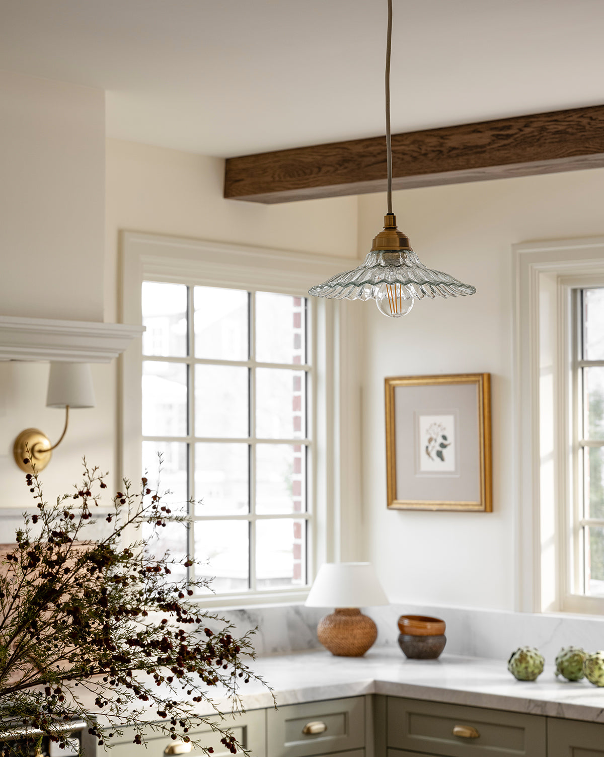 A bright kitchen with a Vendixs x Loeffler Randall Maude Glass Pendant, ruffled glass shade, brass hardware, large windows, green cabinets, marble counters, a framed wall picture, and a vase of dried flowers on the countertop by Vendixs