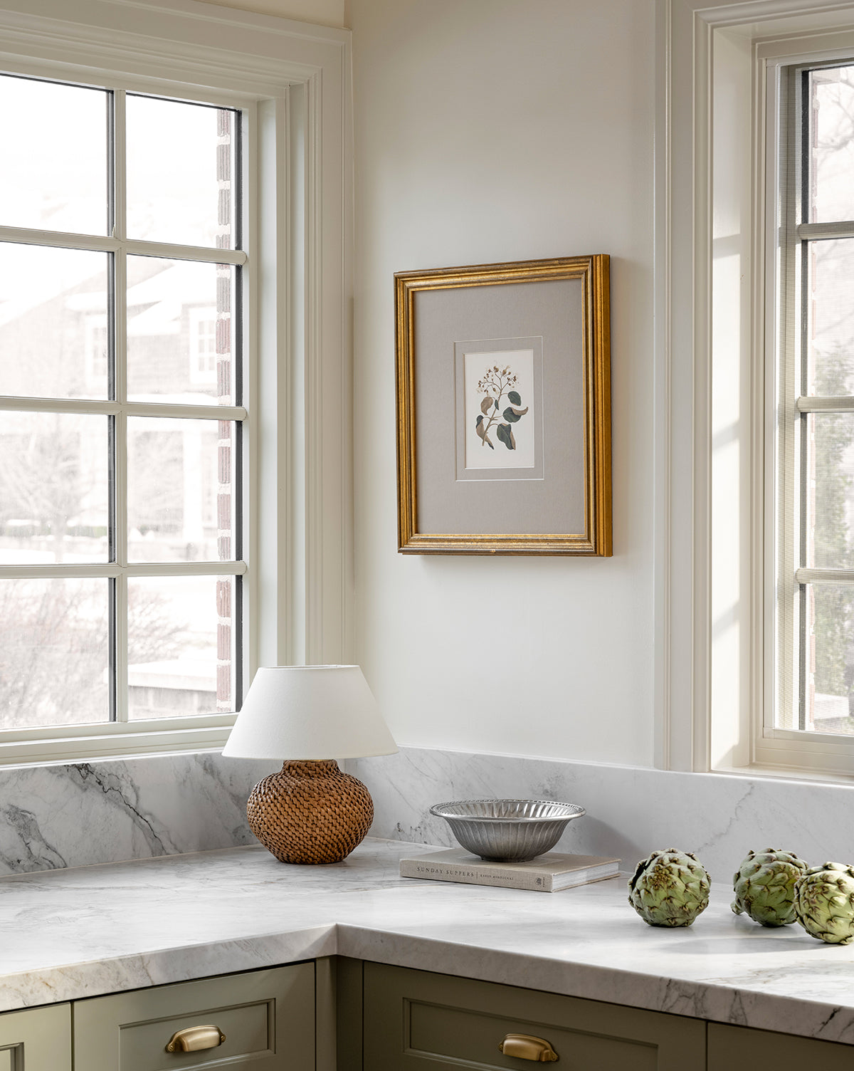 A bright kitchen corner features large windows, a marble countertop, green cabinets, a wicker lamp with a white shade, a silver bowl on books, two artichokes, and the Blossoming Branch vintage wall art in an antique gold frame, by Vendixs