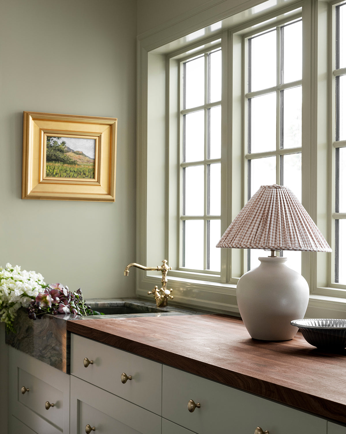 A kitchen with pale green cabinets, brass hardware, and a wood countertop features the Bronwyn Table Lamp with a gingham pleated shade, fresh flowers, and a sink under large windows. A framed landscape painting decorates the wall by Vendixs