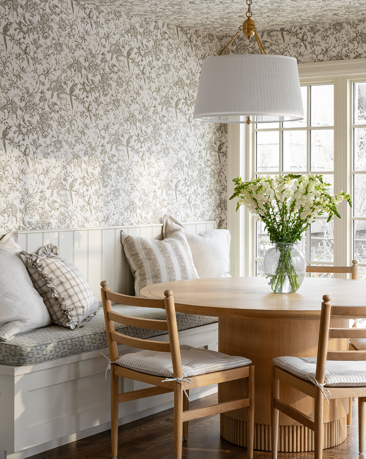 A cozy breakfast nook features the Gilroy Round Oak Dining Table with a reeded base in heritage oak, wooden chairs with cushions, a built-in bench with pillows, floral wallpaper, and large windows that fill the space with natural light by Vendixs