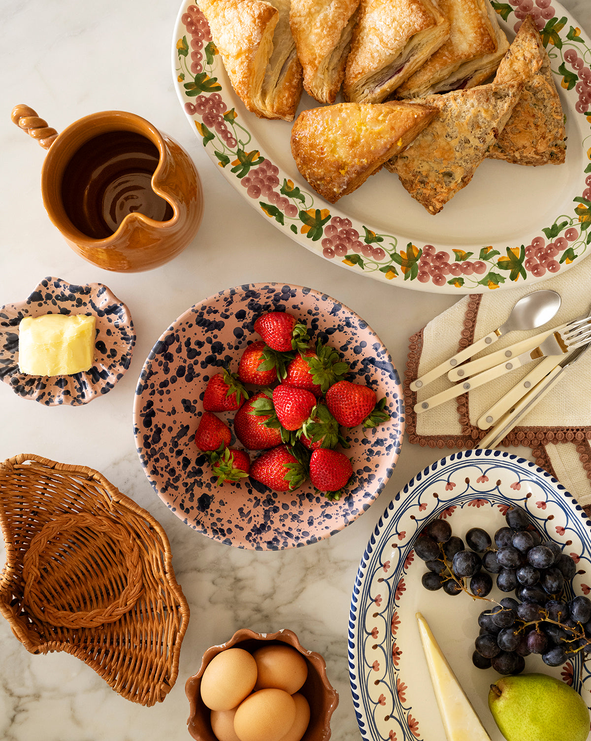 A breakfast table features a floral platter of pastries, fresh fruit, a small dish of butter, the rustic brown Louise Jug with a twisted handle, eggs in a basket, and utensils on a cloth napkin, by Vendixs
