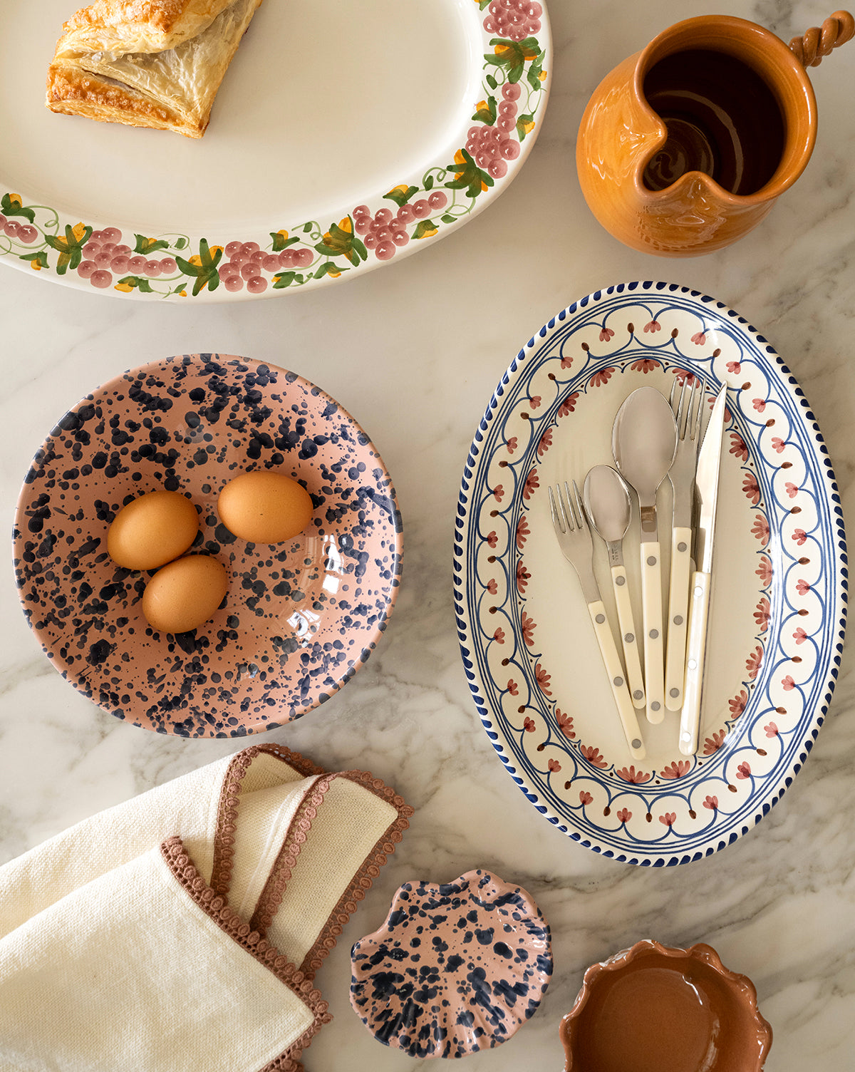 A Splatter Scallop Dish with three brown eggs, an artisan ceramic platter with cutlery, a terracotta cream bowl, a pastry on a floral plate, and folded napkins rest on a marble tabletop, by Vendixs