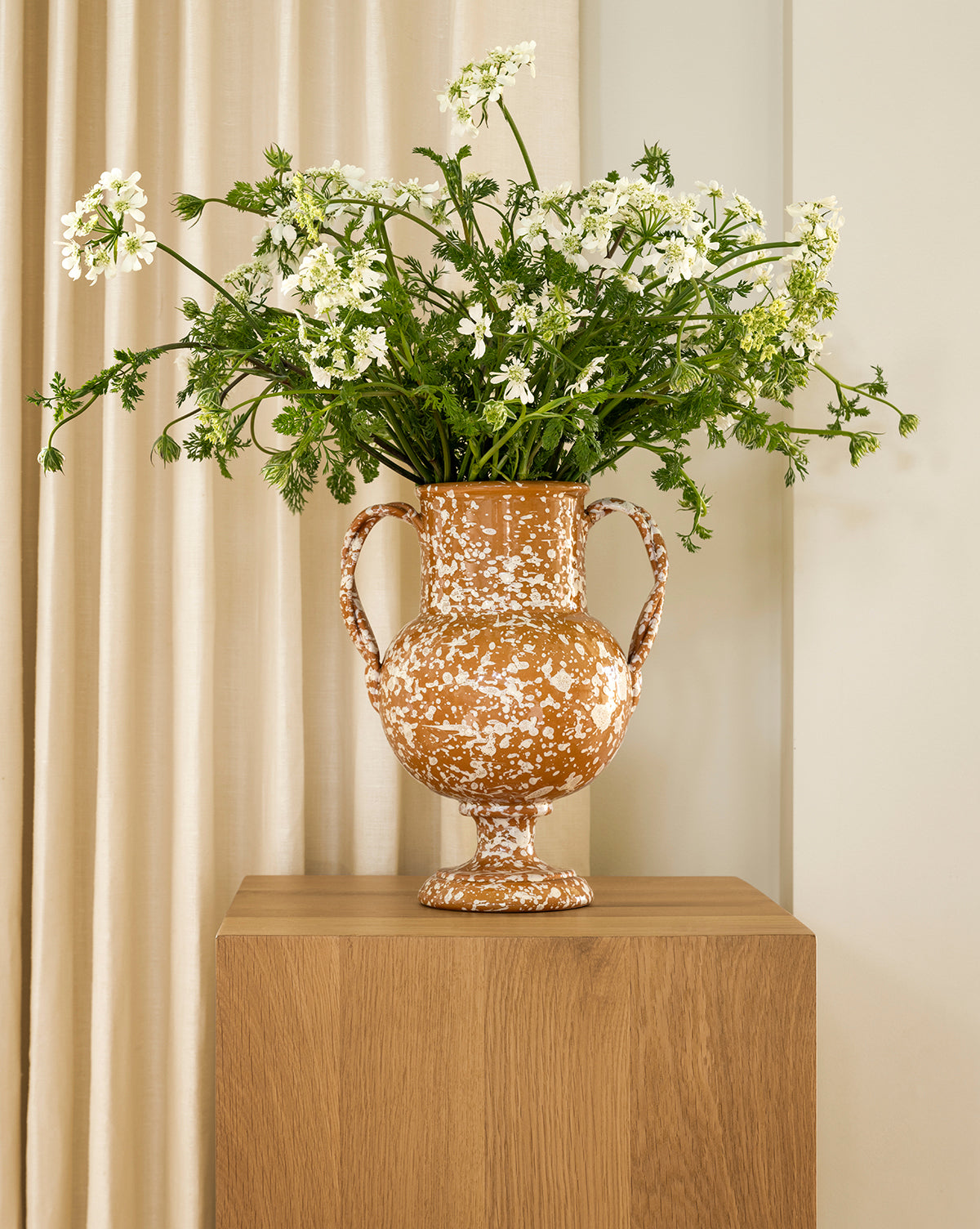 The Splatter Verona Vase, featuring two handles and a speckled design, holds white flowers and green foliage on a wooden pedestal against neutral beige curtains, by Vendixs