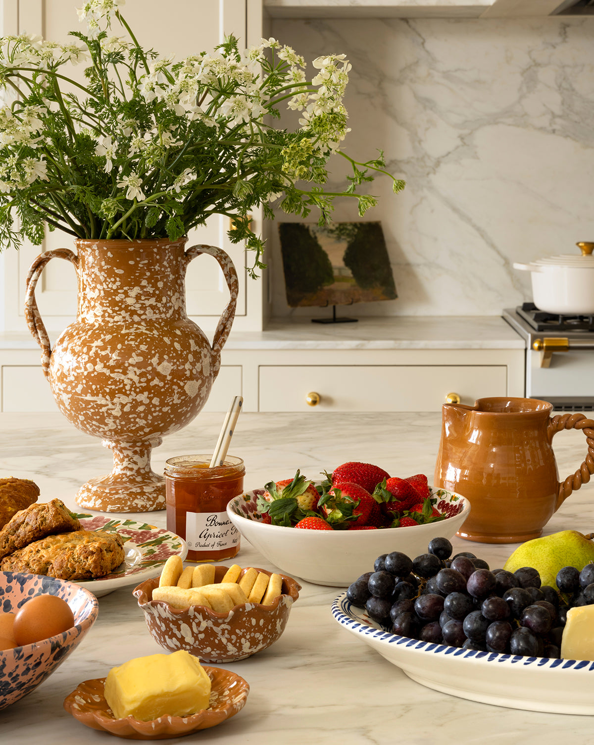 A kitchen counter displays the Splatter Verona Vase filled with white wildflowers, surrounded by strawberries, grapes, jam, pottery mugs, scones, butter, eggs, pears, and cookies—all beautifully arranged for breakfast, by Vendixs