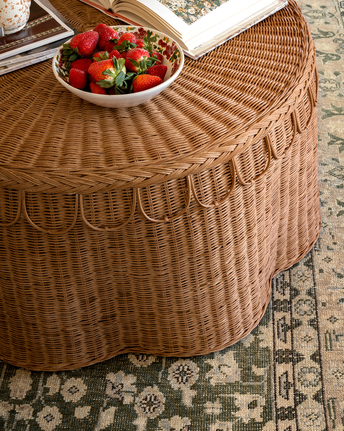 A round wicker table with a scalloped edge holds the Vine Large Bowl filled with strawberries and an open book, set on a green and beige patterned rug, by Vendixs
