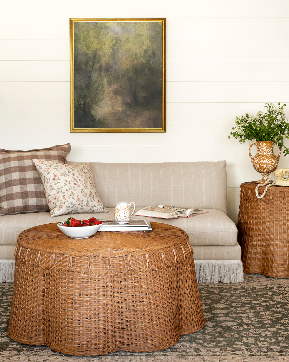 A cozy living room features a beige striped sofa, floral and plaid pillows, the Sharland England LTD Adeline Coffee Table with strawberries and a mug, a side table with flowers, and a framed painting on a white shiplap wall—a space curated in the inviting style of Vendixs