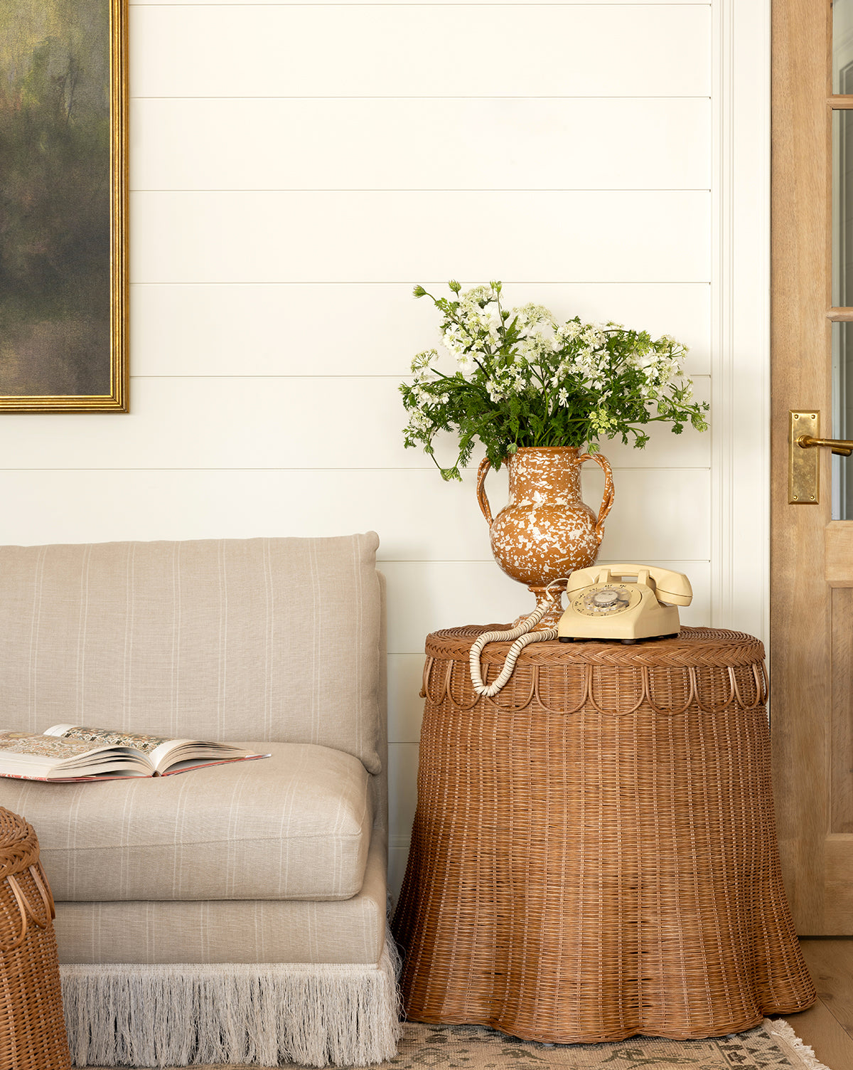 A cozy living room corner with a beige loveseat, an open book, and the Adeline Side Table by Sharland England LTD topped with a yellow rotary phone and white flowers, beside a wooden door and a painting on a white shiplap wall, Vendixs