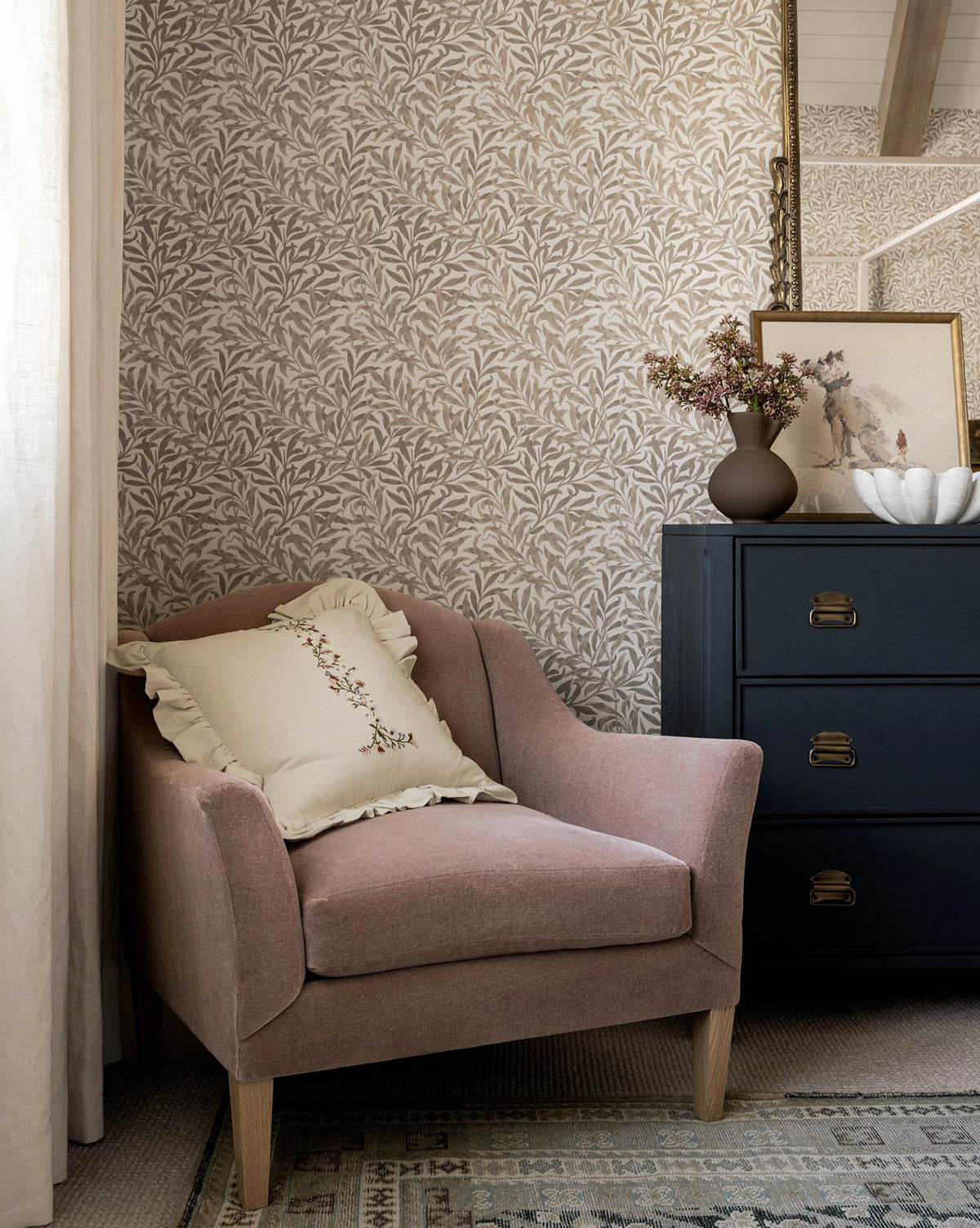 A cozy corner features the classic silhouette of a blush Makerspalm Prudence Lounge Chair with a ruffled, embroidered pillow beside a dark dresser topped with framed art, a ceramic bowl, dried flowers, and subtle leafy wallpaper—McGee & Co.