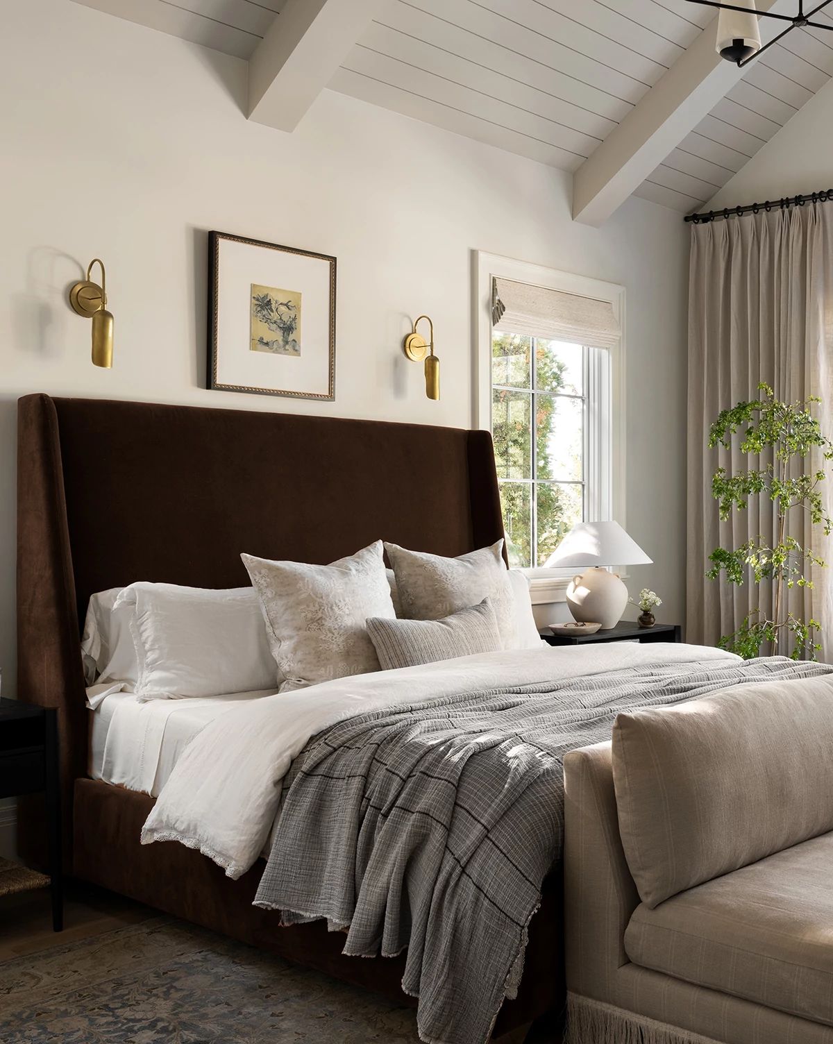 A bedroom featuring the Walt Bed Tall Headboard in dark brown with solid oak legs, white bedding, neutral pillows, gray blanket, two wall sconces, framed art, window curtains, and a leafy plant beside the bed by McGee & Co.