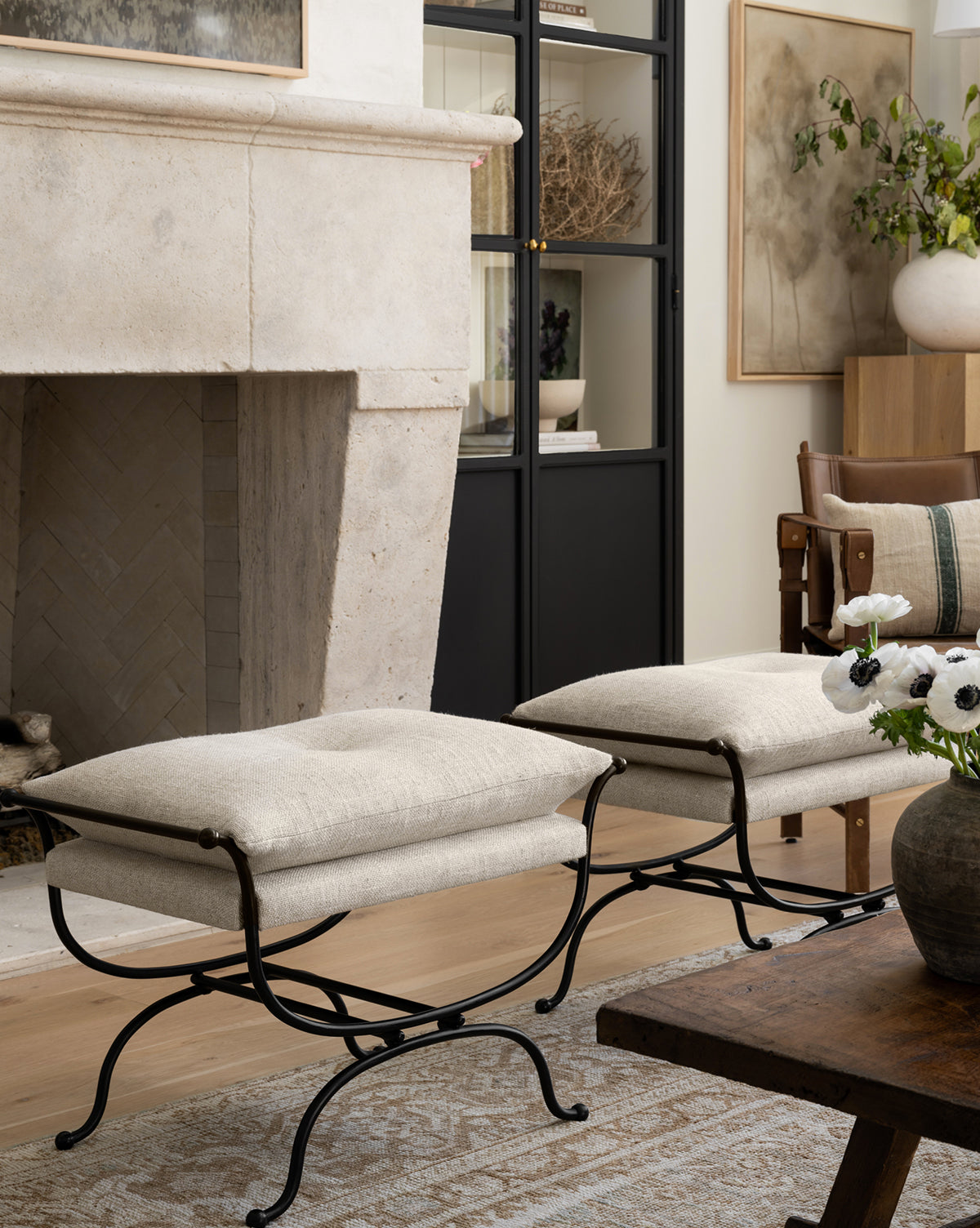 Two Makerspalm Alameda Stools with linen upholstery and matte black iron bases are placed in front of a stone fireplace in a cozy living room, accompanied by a wooden coffee table, vase of flowers, and decorative shelves. Vendixs