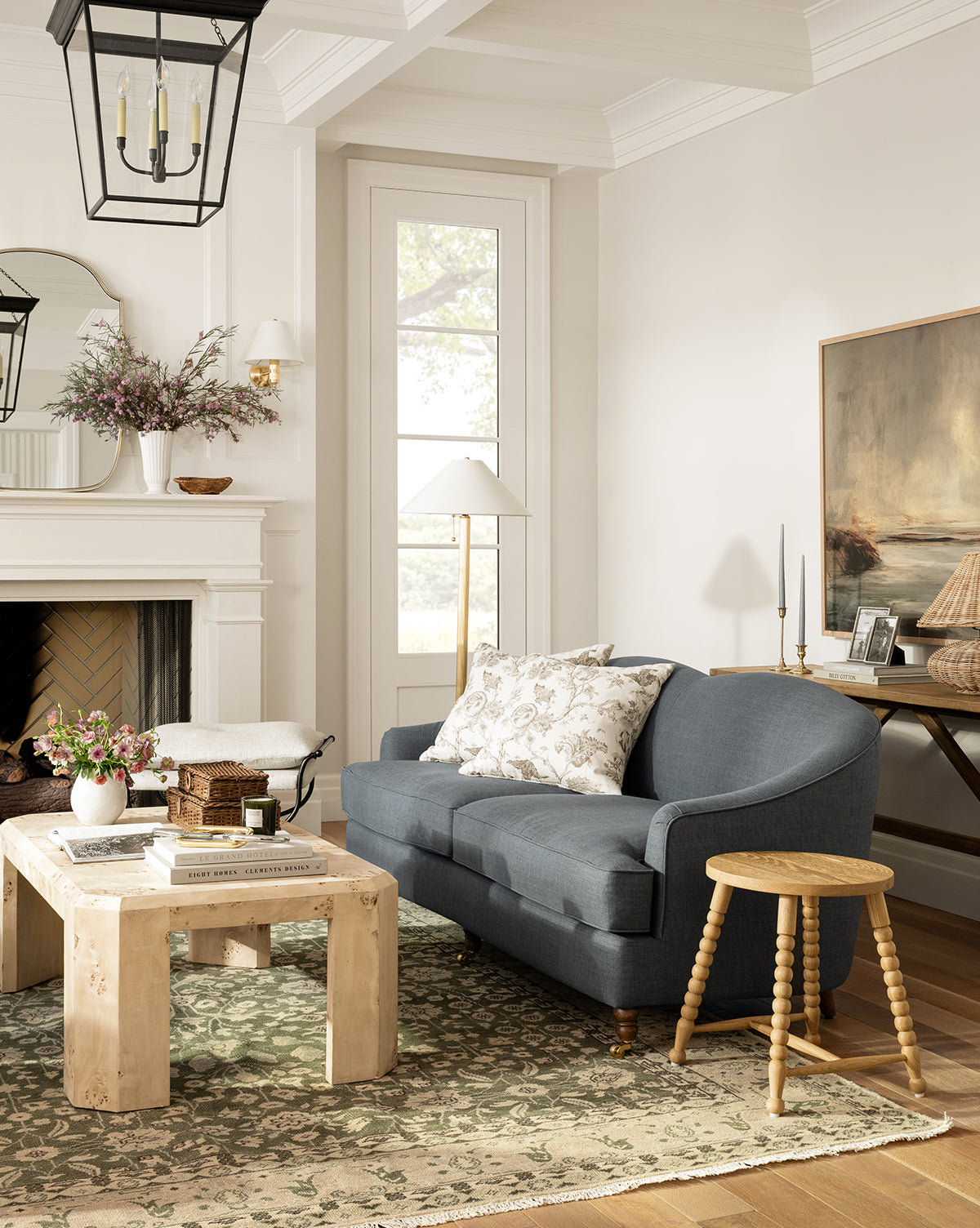 A cozy living room with a blue sofa, floral pillows, wooden coffee table, and the Fernwood Hand-Knotted Wool Rug in green tones. A fireplace topped with flowers and large windows complete the scene alongside decorative accents and artwork by Vendixs