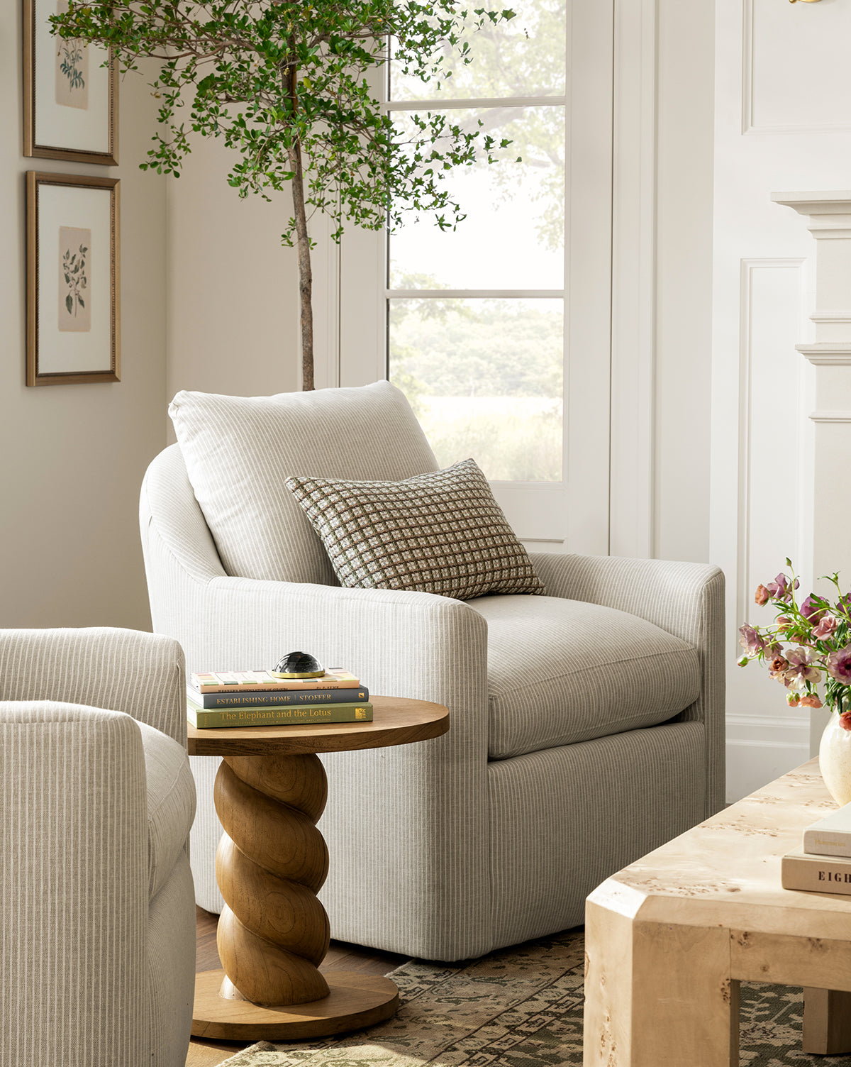 A cozy living room corner features a Rowe Fine Furniture Quinnton Swivel Chair in light gray, paired with a patterned cushion, round wood side table, books, potted plant, and framed art on a cream wall with natural light from the window—McGee & Co.