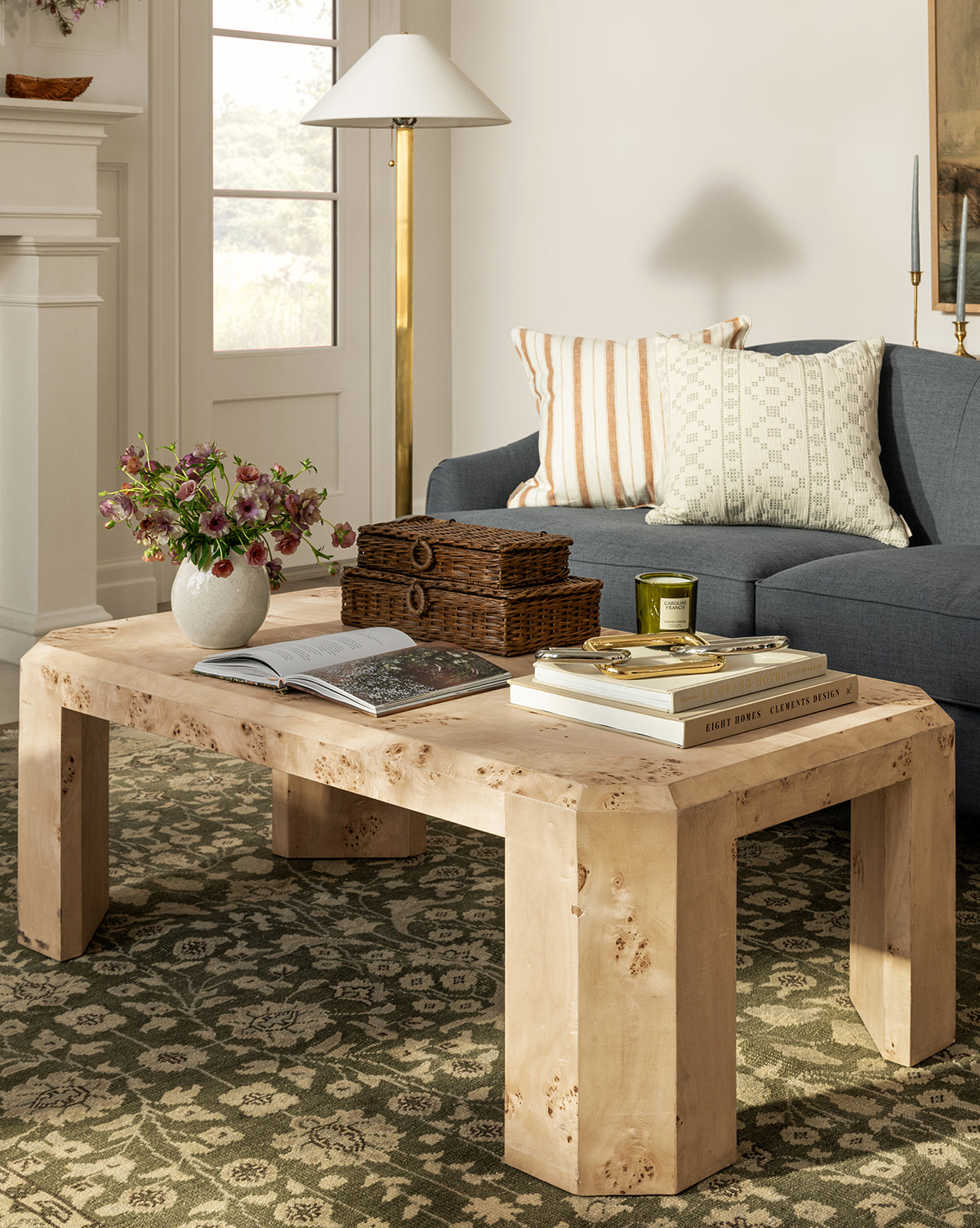 The Rocio Burl Coffee Table holds books, a vase of flowers, a candle, and wicker boxes in a cozy living room with a blue sofa, patterned cushions, a floor lamp, and a patterned rug covering the floor, by Vendixs