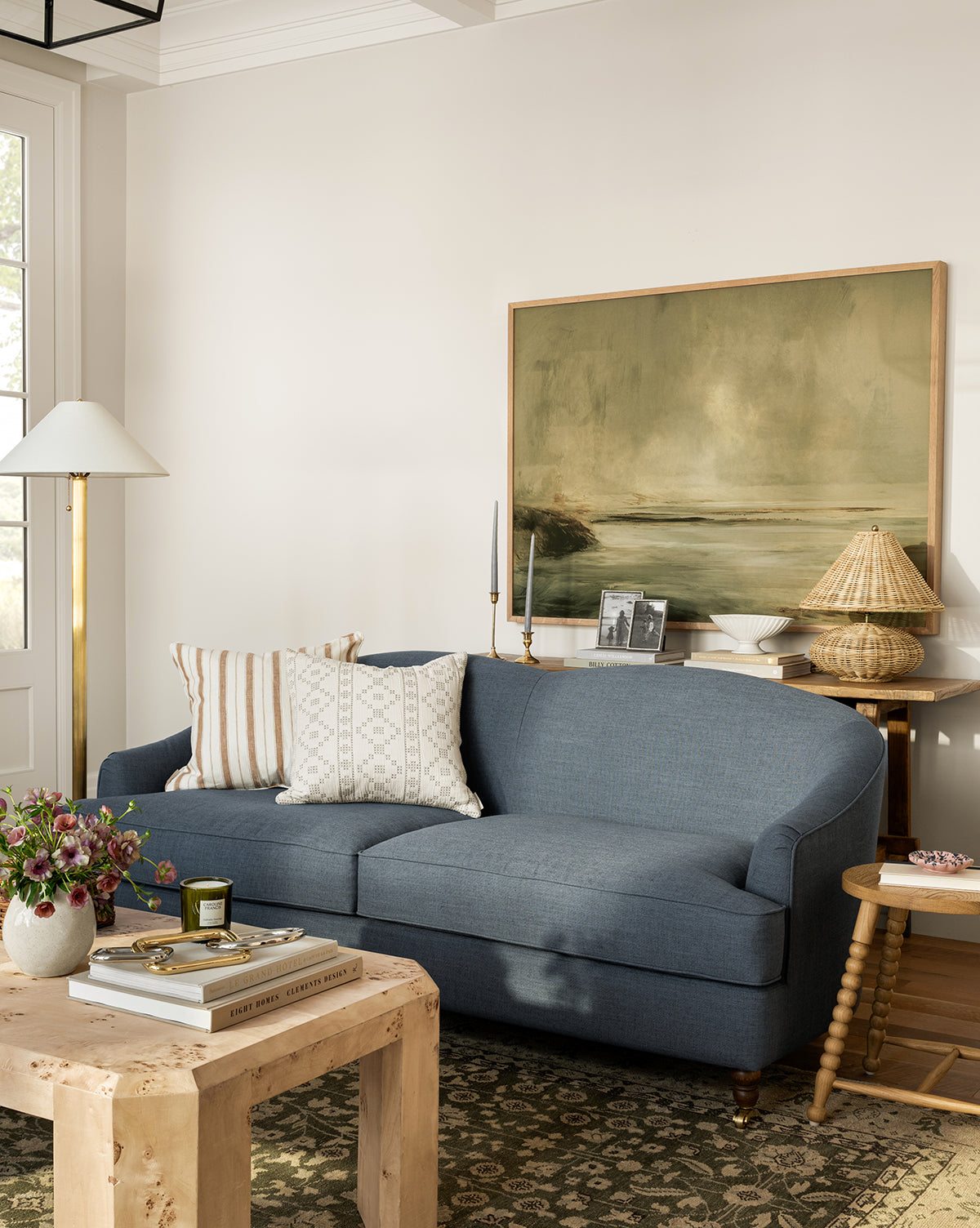 A cozy living room with a blue sofa, patterned pillows, a wood side table and woven lamp, a tall floor lamp, and the Misty Blue Headland gallery-wrapped landscape canvas above a coffee table styled with books and flowers, by Vendixs