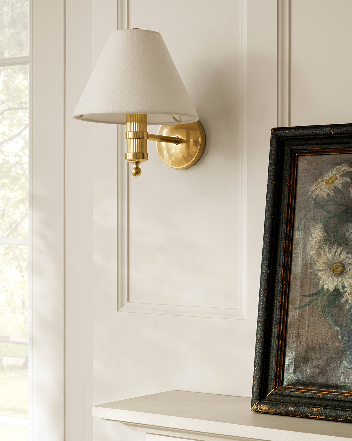 A Meredith (China) Ltd Townes Sconce with a linen shade is mounted on a white paneled wall above the mantel, beside a framed floral painting. Sunlight pours in from the window to the left, Vendixs