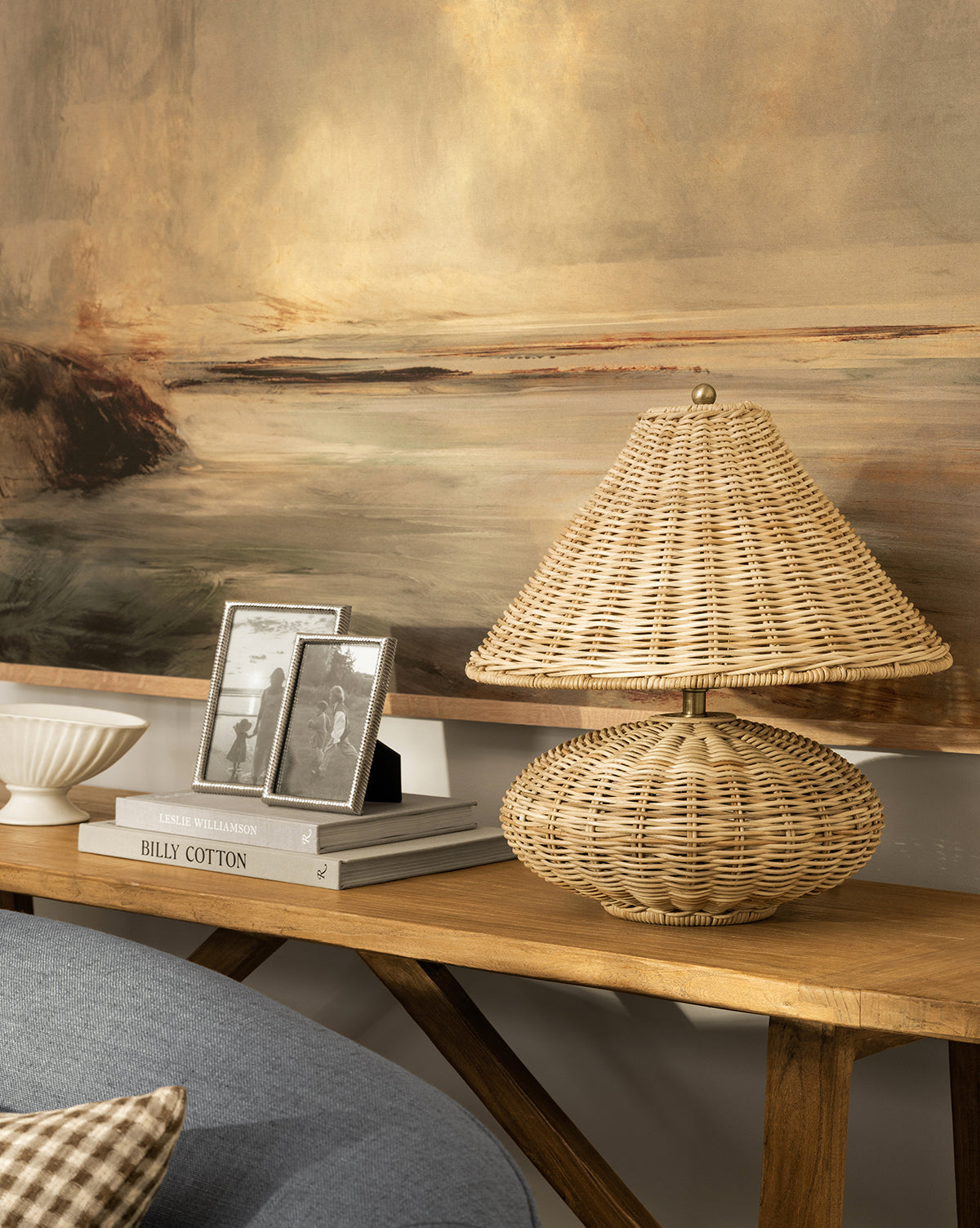 The Palmetto Table Lamp with a woven shade sits on a wooden console table beside framed black-and-white photos, a white bowl, and books, with a large landscape painting behind—perfect for organic home decor, by Vendixs
