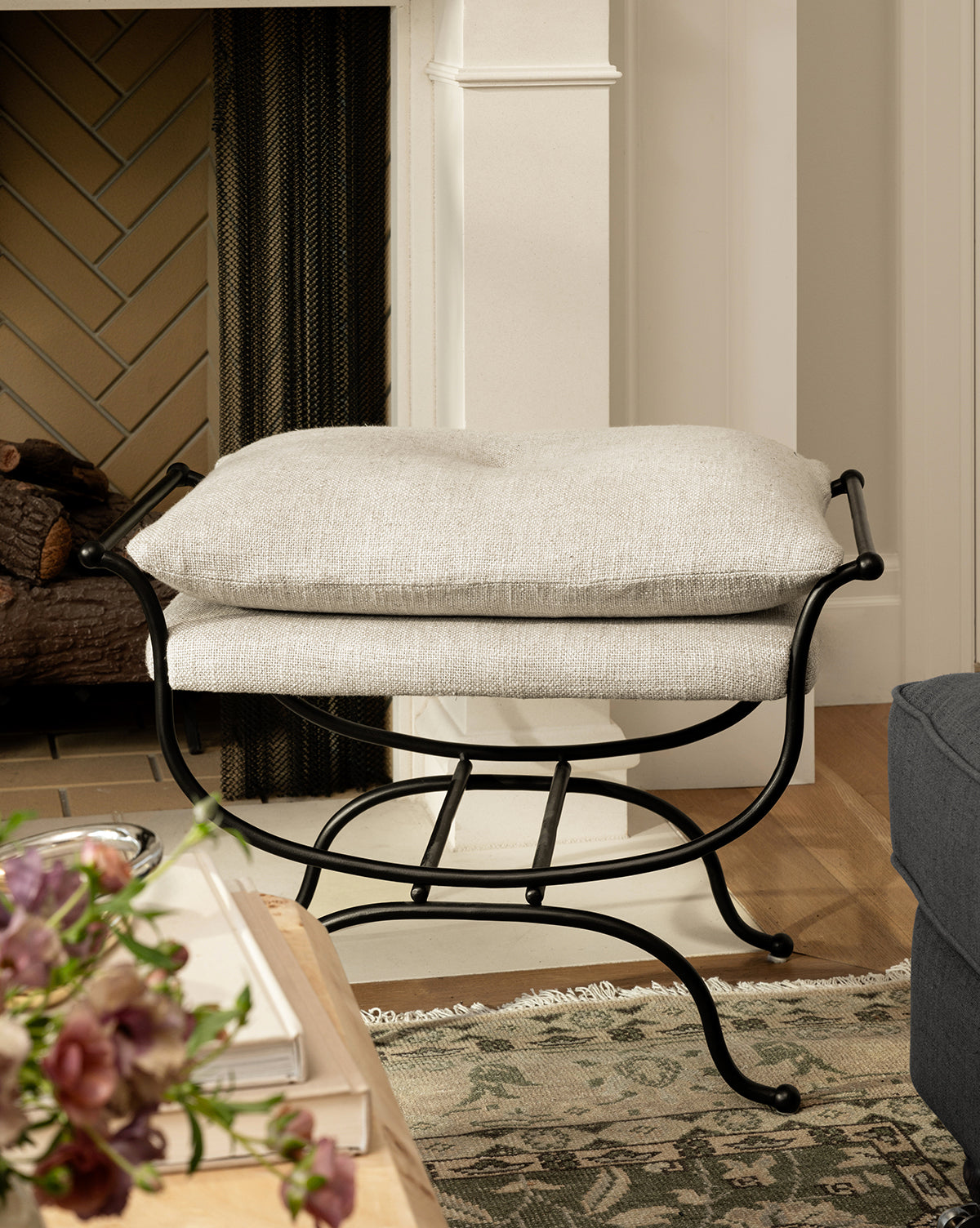 A cozy living room showcases the Makerspalm Alameda Stool with a matte black iron base and linen upholstery by the fireplace, next to a blue sofa and wooden table with flowers on a patterned rug, Vendixs