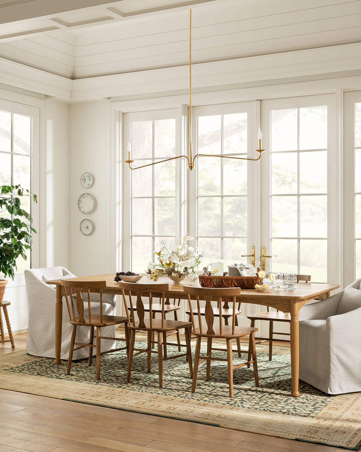 A bright dining room features the handcrafted Phipps Dining Table set for a meal, six wooden chairs, two white armchairs, a brass modern chandelier, large windows, wall plates, a potted plant, and a patterned rug on wooden floors, by Vendixs