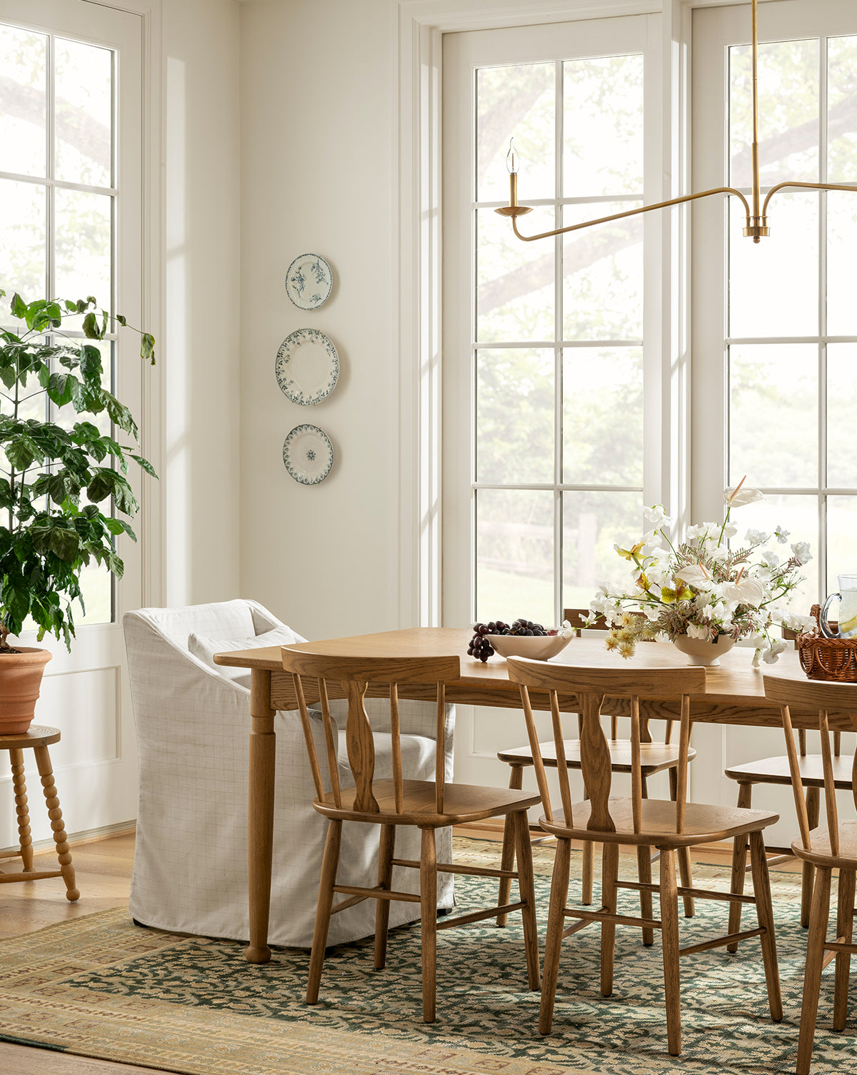 A bright dining space with large windows, a solid oak table, mismatched chairs including the Whittaker Chair by Yang Chen, with a potted plant, and floral centerpiece. Light fills the cozy room. Vendixs