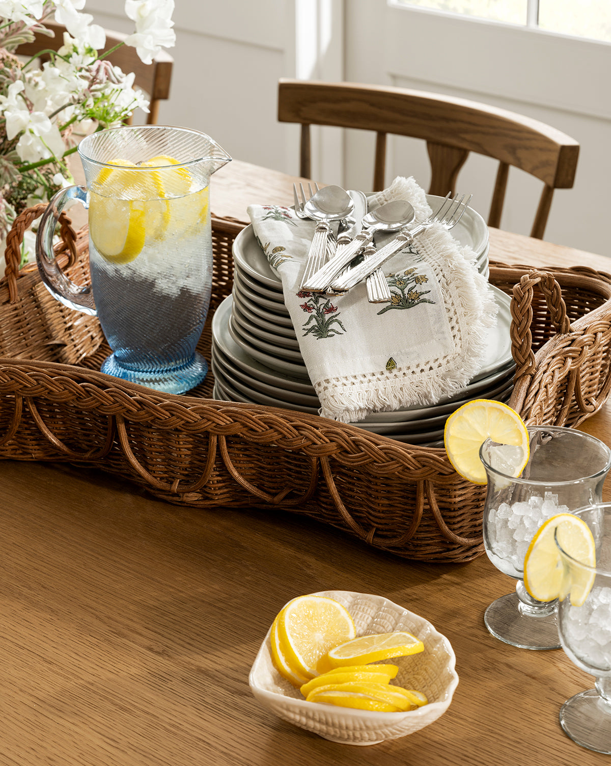 A wicker tray holds the Spiral Jug filled with lemon water, stacked plates, and cutlery with napkins. Handcrafted glass tumblers with lemon slices and a bowl of lemon wedges sit nearby, all bathed in bright natural light by Vendixs