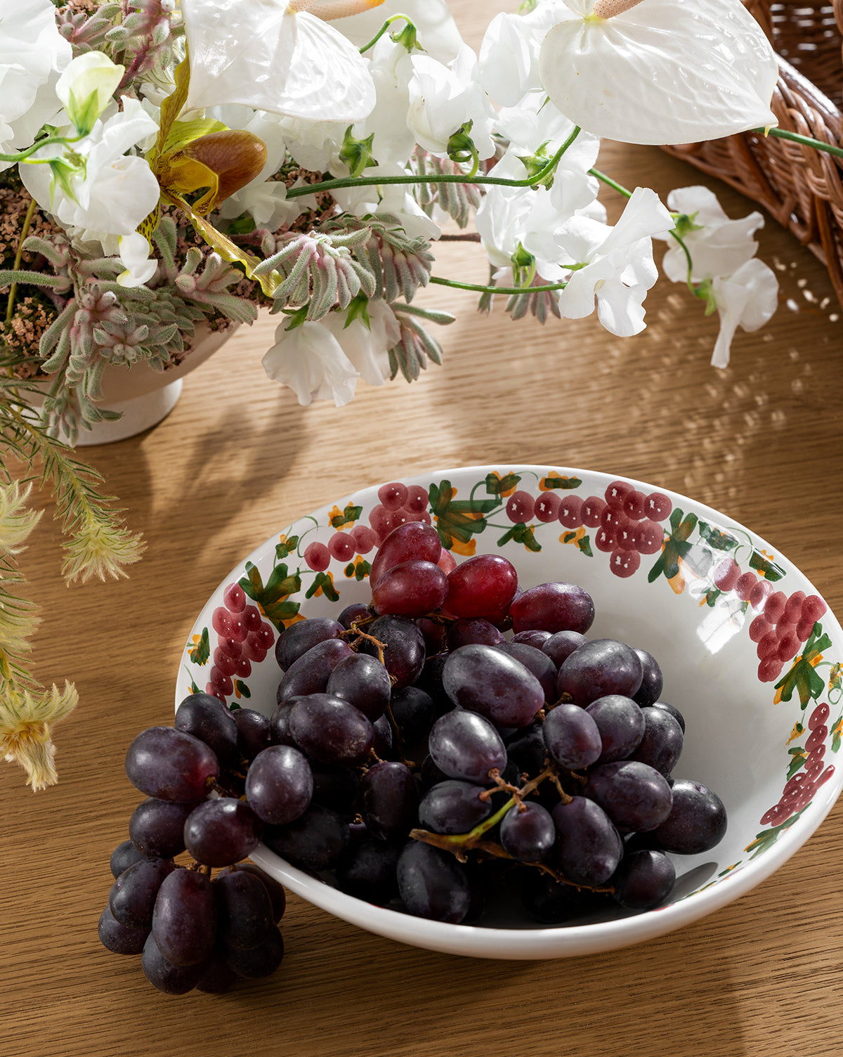 The Vine Large Bowl, a handmade ceramic bowl with a grapevine motif, holds bunches of purple grapes on a wooden table. A white floral arrangement and part of a wicker basket add to the charming scene by Vendixs