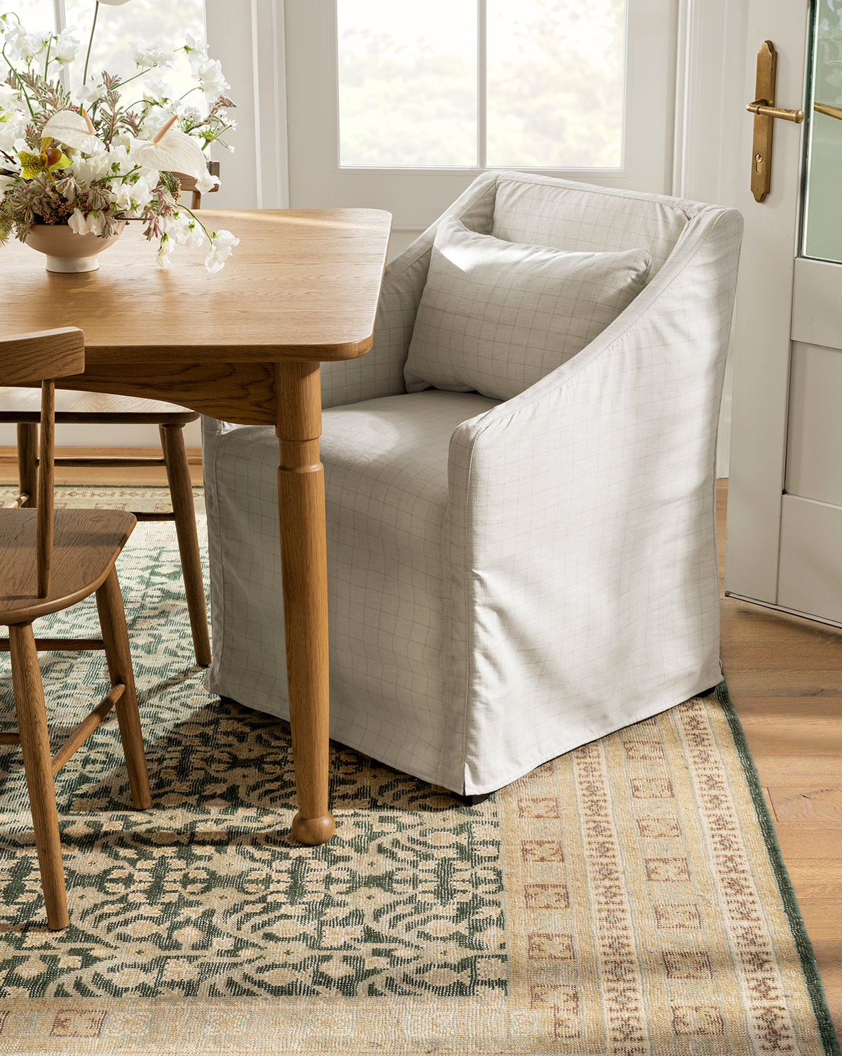 An Olivier Slipcover Dining Arm Chair by Rowe Fine Furniture, Inc. in light gray with a feather and down cushion and matching pillow sits at a wooden dining table beside another chair, atop a patterned rug, with floral accents and natural light nearby. Vendixs