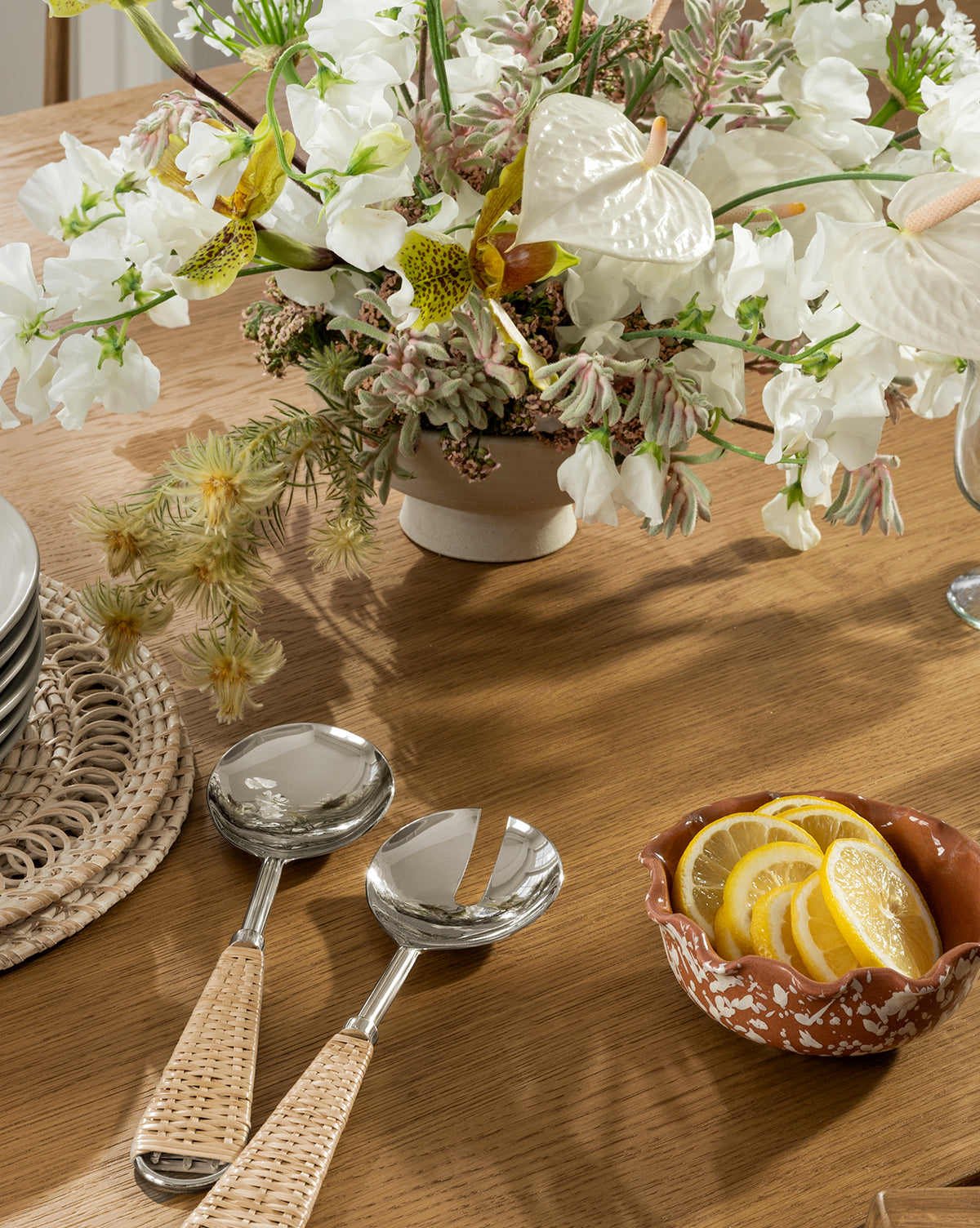 A wooden table with a ceramic vase of white flowers, two woven-handled serving utensils, a basket of stacked plates, and the Splatter Scallop Dish filled with lemon slices, by Vendixs
