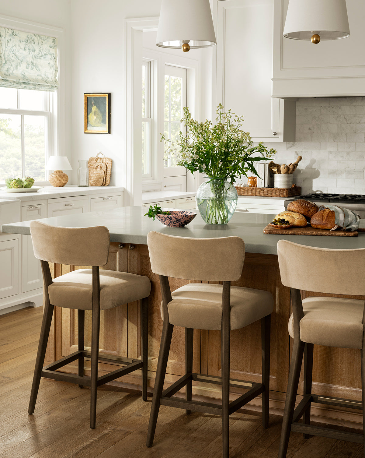 Bright kitchen with a wooden island, three Makerspalm Etienne Stools (Ready to Ship), glass vase with flowers, bread and fruit on the counter, white cabinets, and large windows letting in natural light, Vendixs