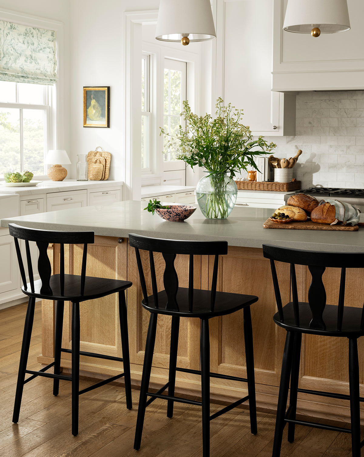 A bright, modern kitchen with a light wood island features three Whittaker Counter Stools by YANG CHENG WOODEN INDUSTRIES INTERNATIONAL (VIETNAM) CO LTD, each with a spindle back, plus fresh bread and flowers on the counter for an airy vibe—finished with design touches inspired by Vendixs